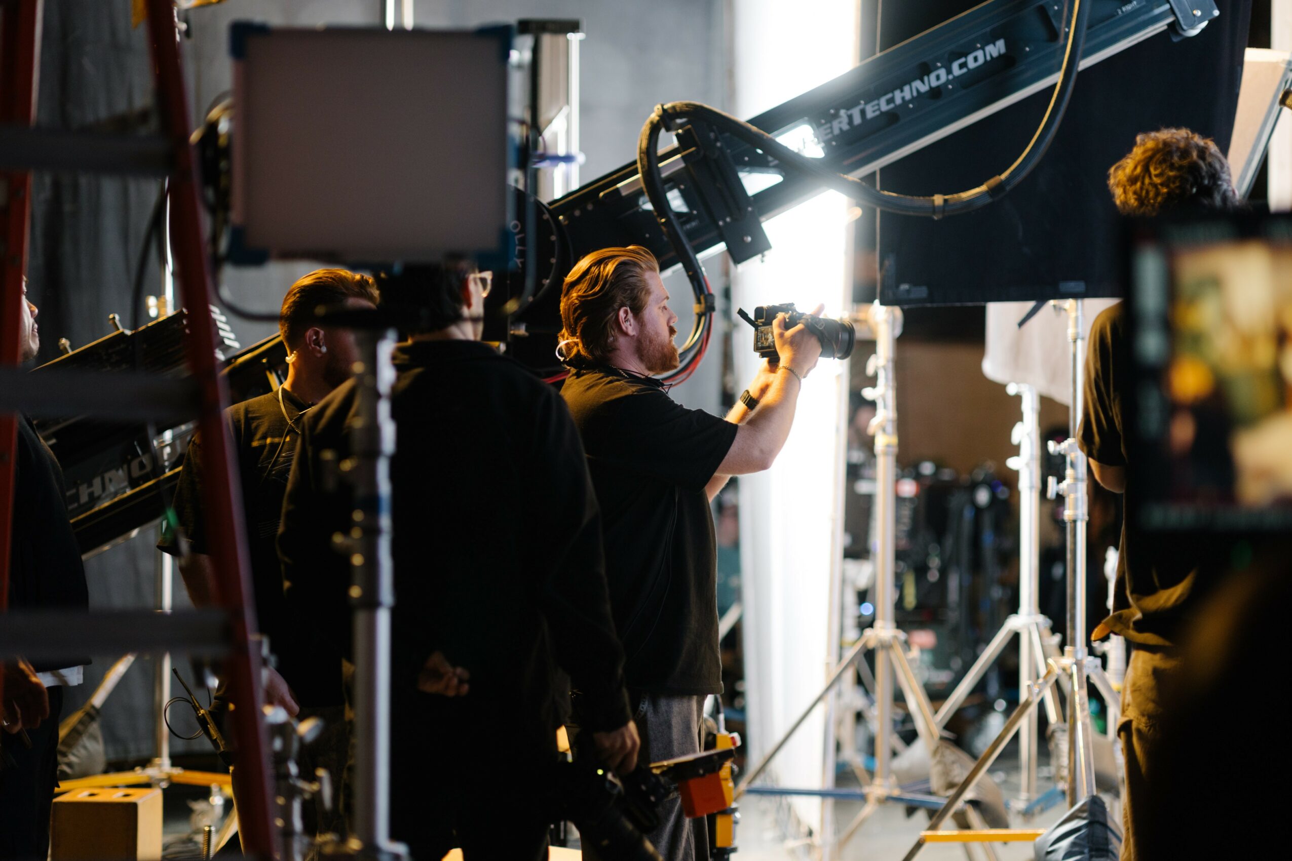 Filmmaker adjusting equipment on set with crew members and lighting in background.