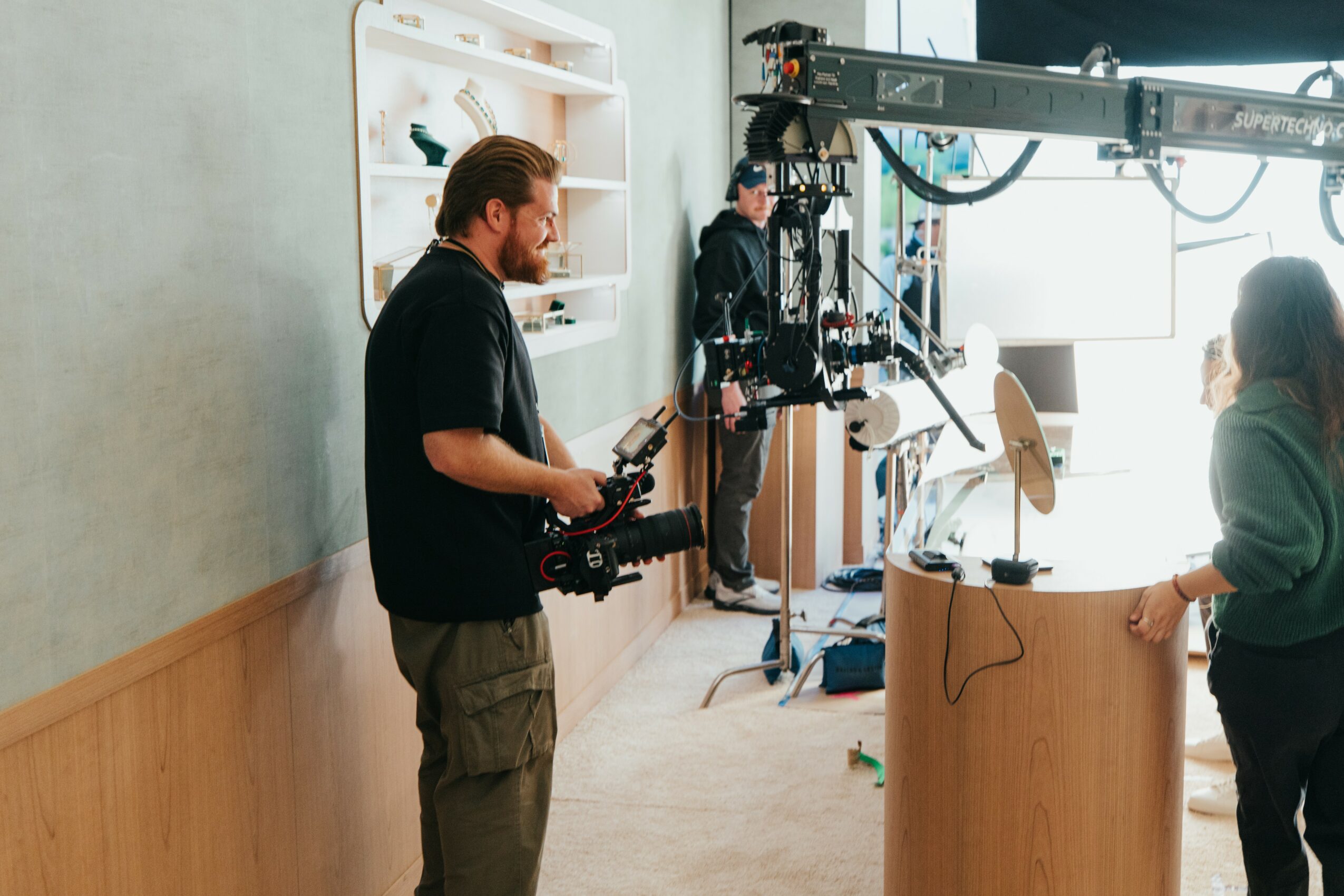 Two people operating camera equipment in a room with a person standing at a podium, bright natural light coming through windows.