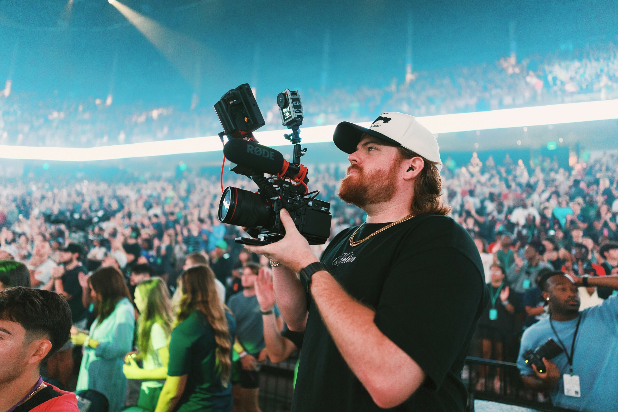 A man with a beard wearing a white cap and black shirt holding a camera with a large lens at a crowded event.
