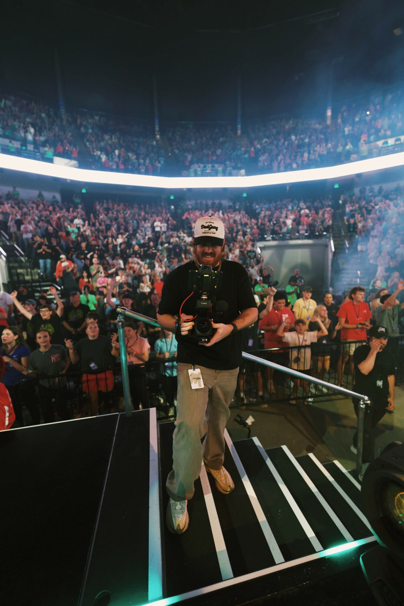 Person standing on stage with a large crowd in the background, stage lights, and smoke effects, holding a microphone.