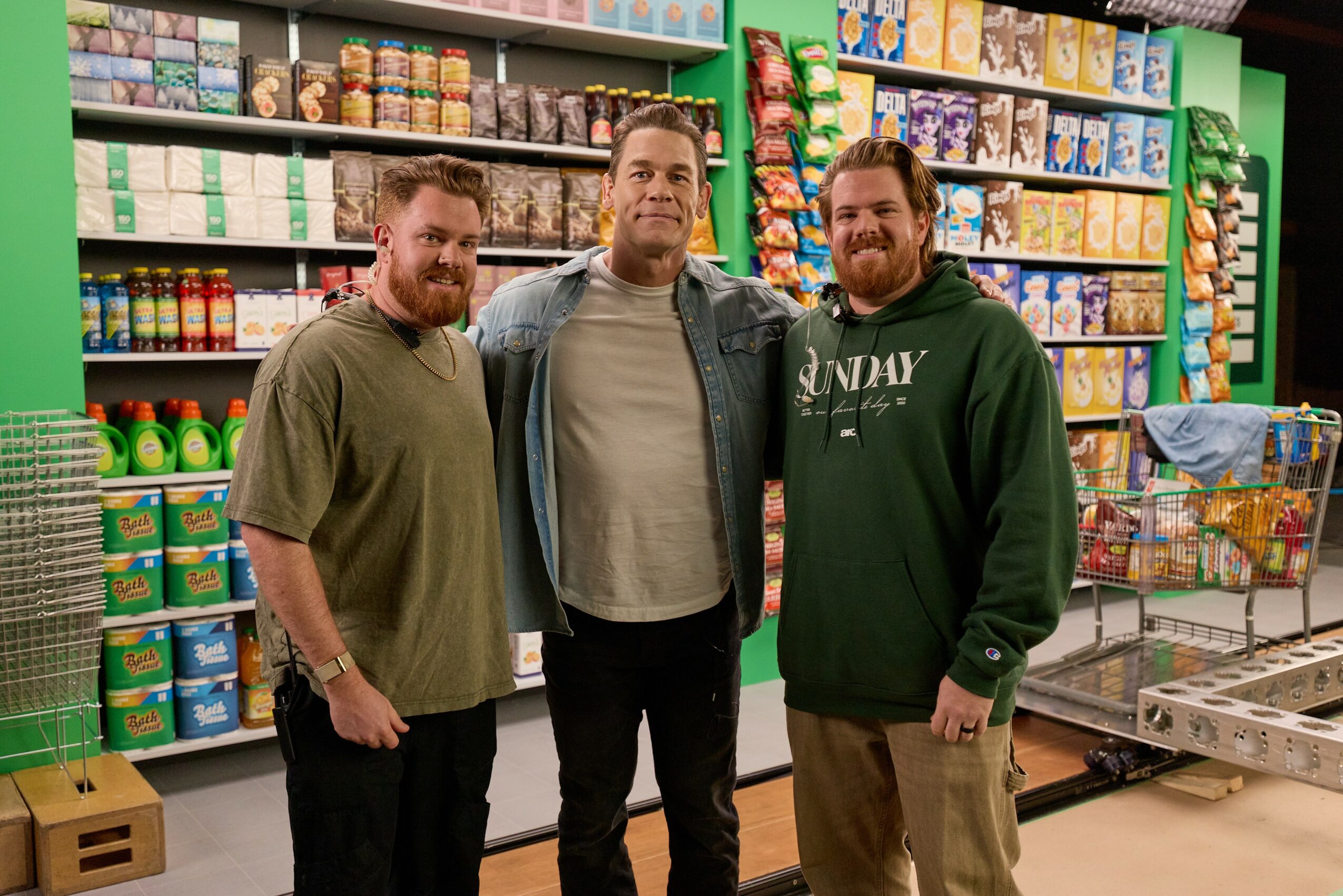 Three men stand together in a grocery store aisle with shelves of canned goods and snacks behind them.