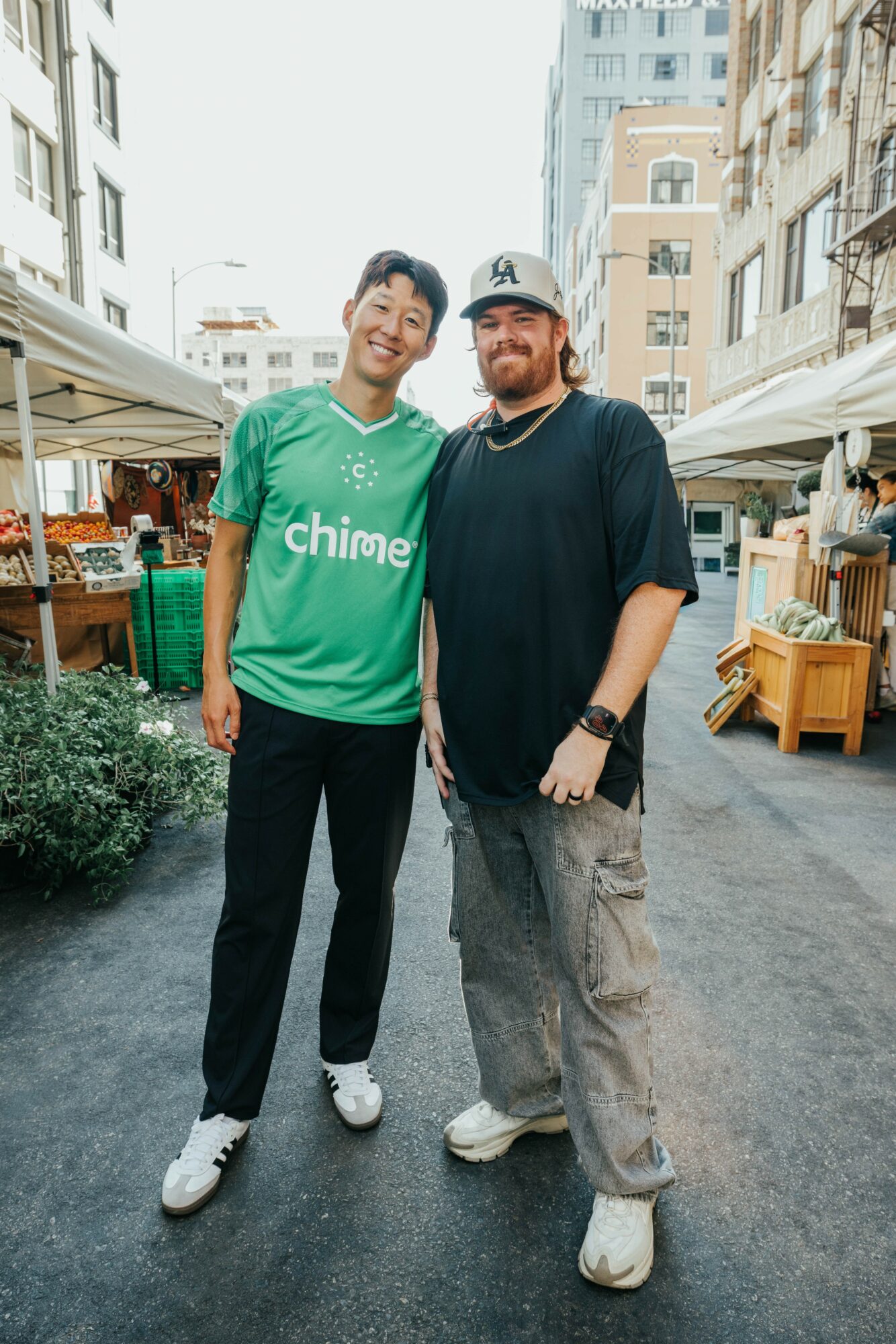 Two men standing together outdoors in an urban market area, smiling, with tents and buildings in the background.