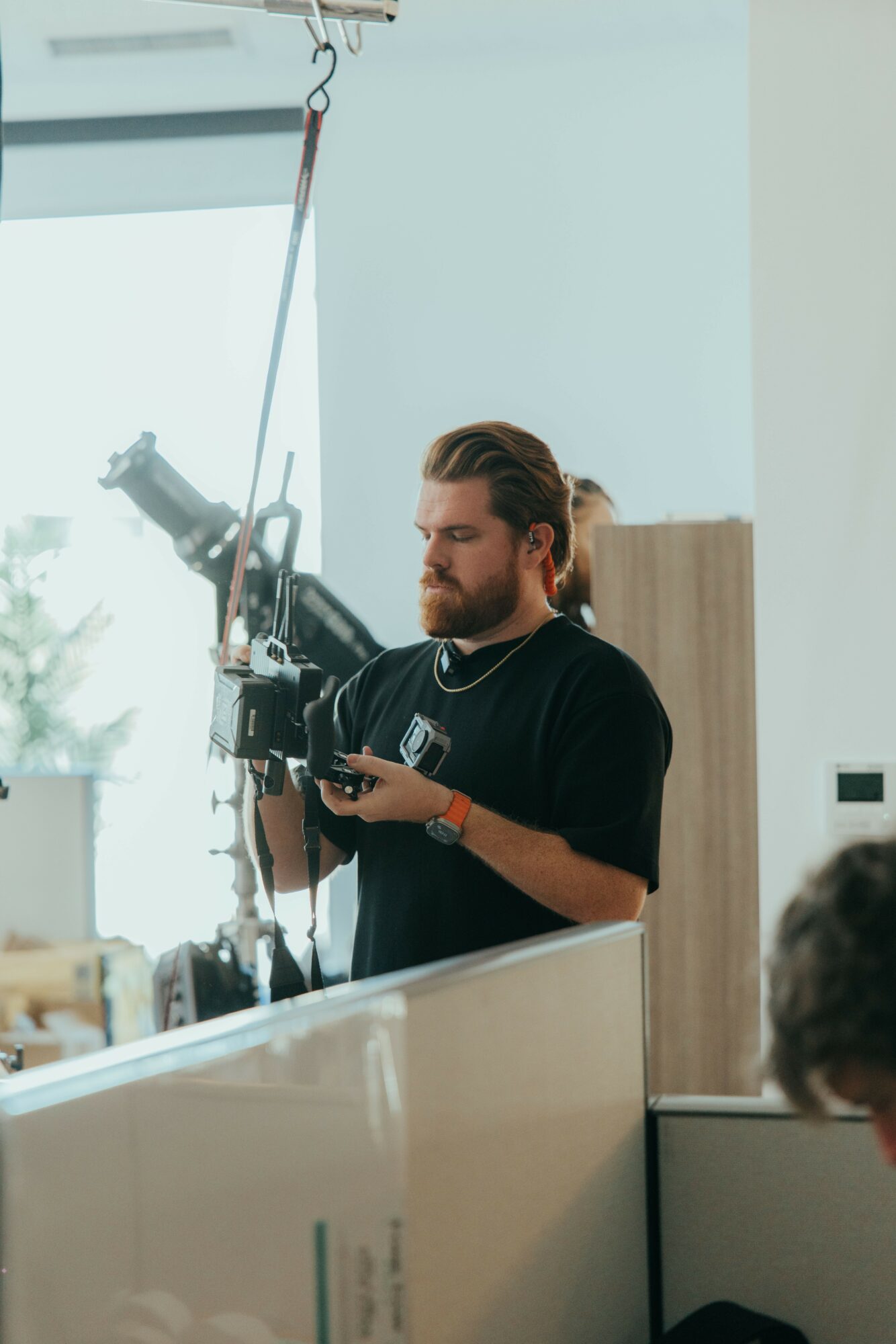 Man with long hair and beard operating a camera indoors, with a window in the background.