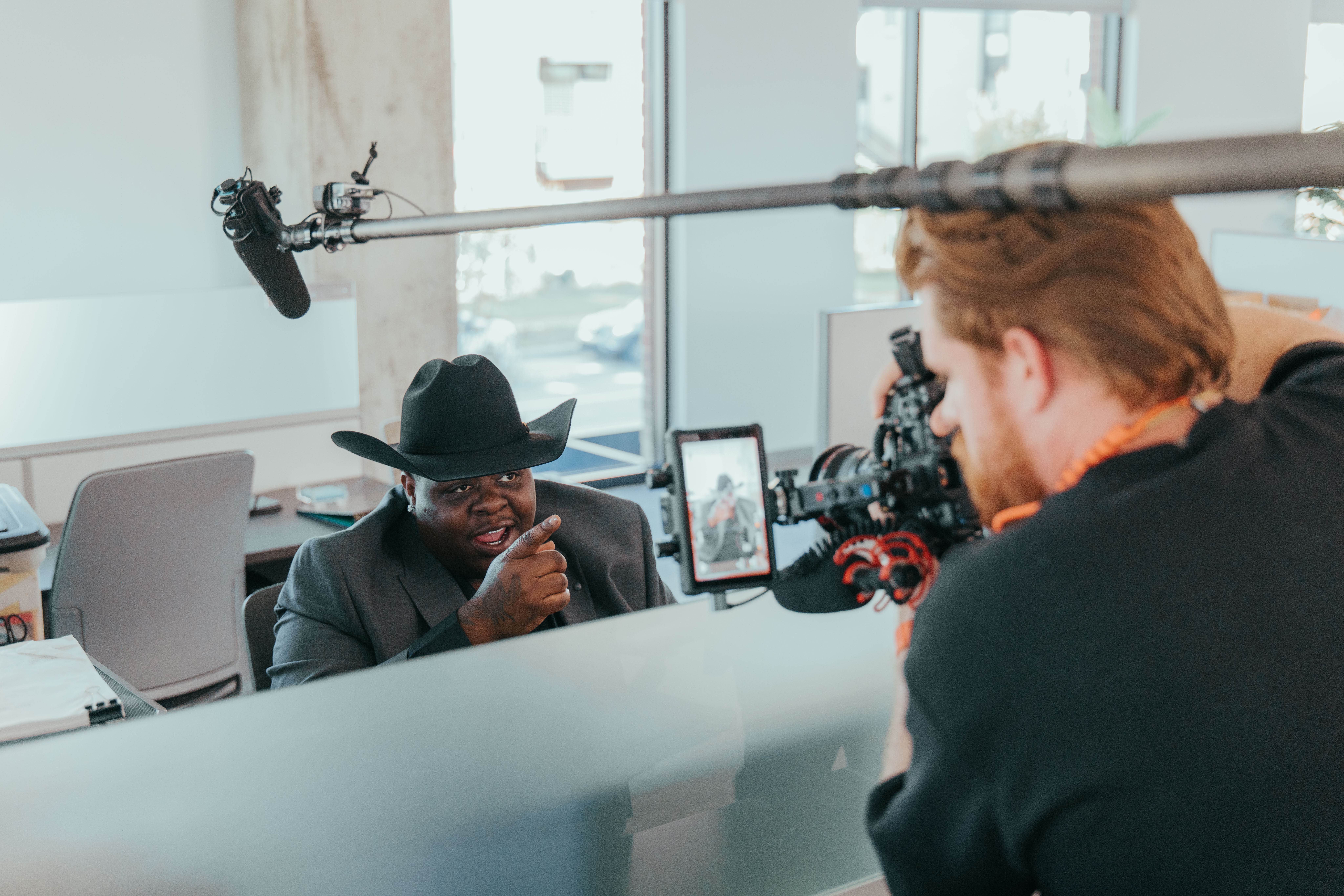 Person in a suit and hat being interviewed in a modern office, with a camera and crew member visible.