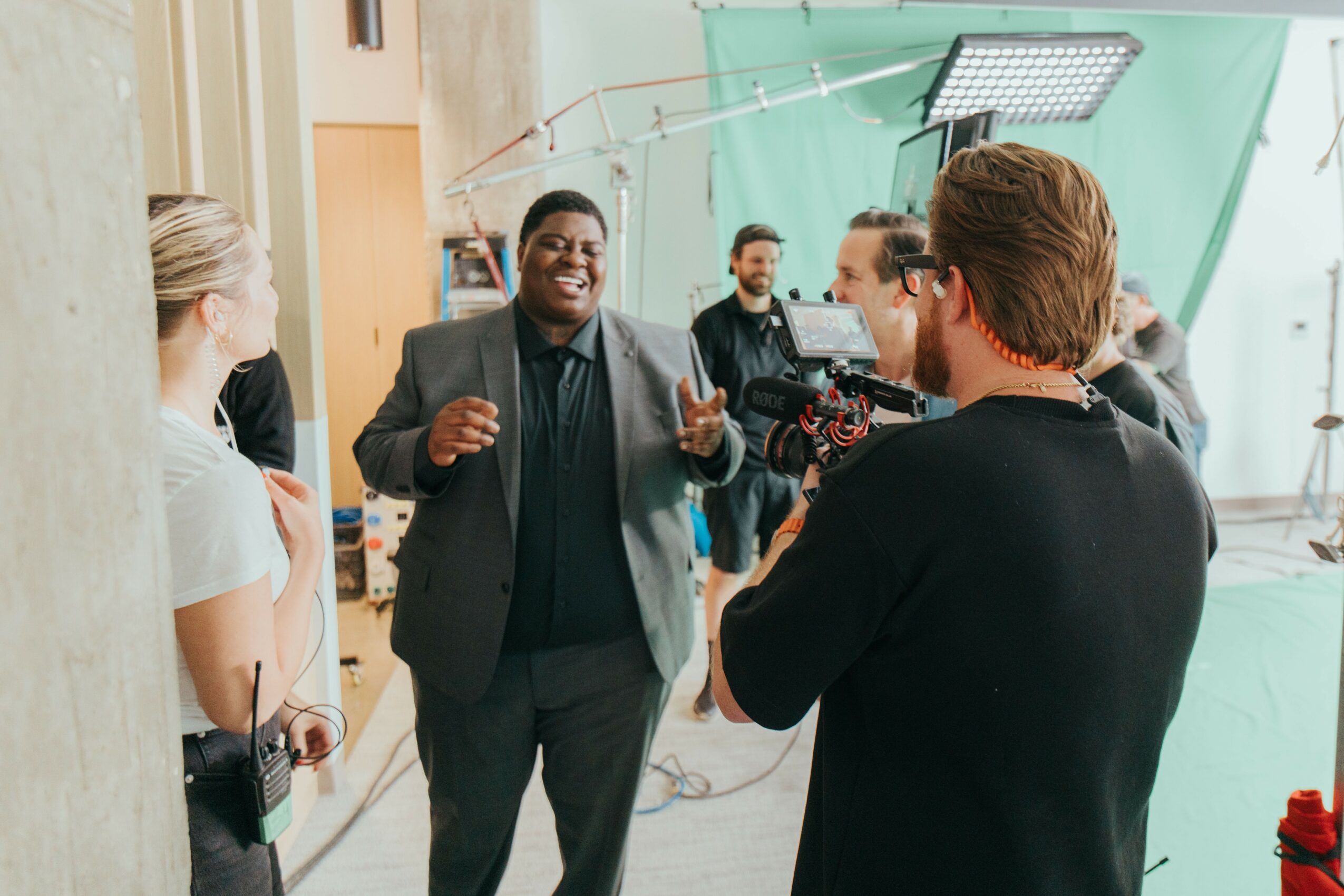 Group of people in a studio, with a man speaking and a camera operator filming him, surrounded by lighting equipment.