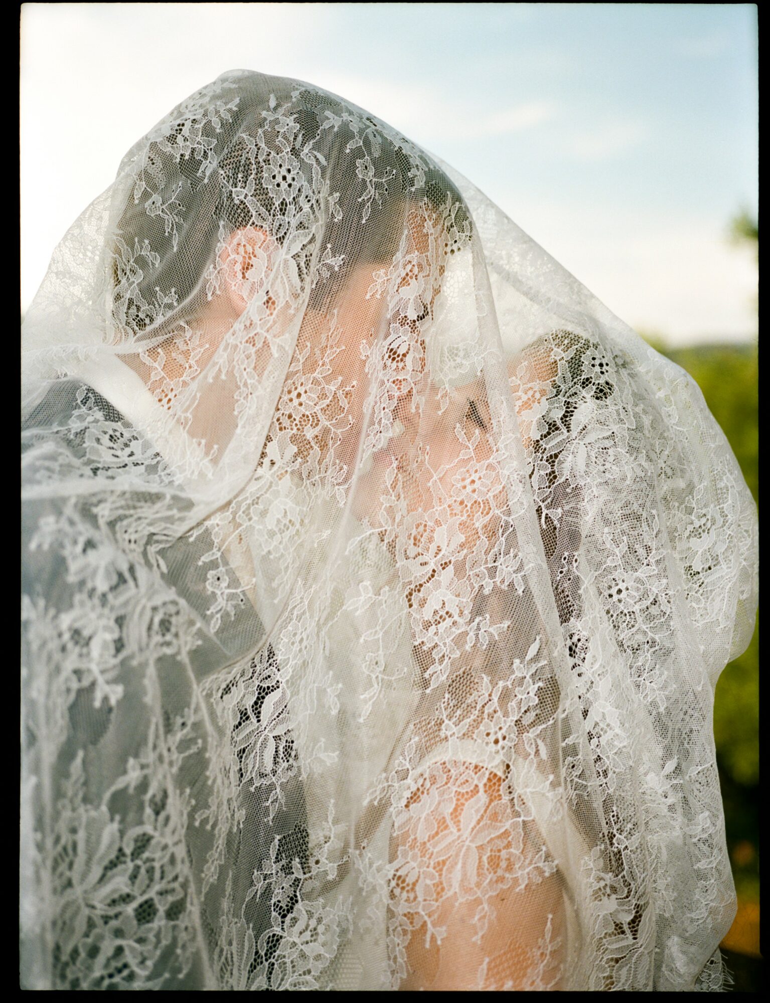 Two people face each other under a lace veil outdoors, with a blue sky and greenery in the background.