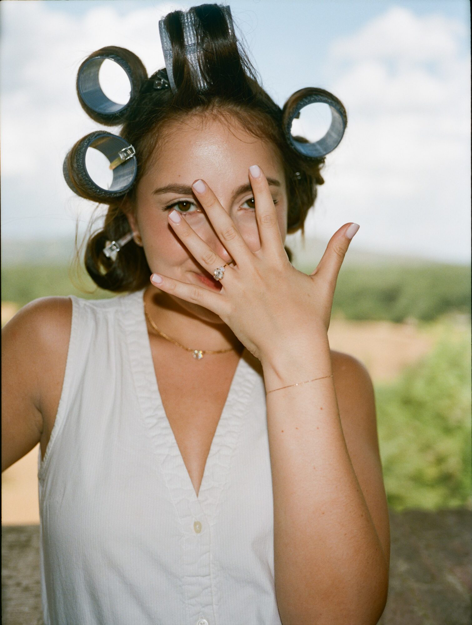 Woman with curlers in her hair, covering part of her face with her hand, outdoors with a cloudy sky background.