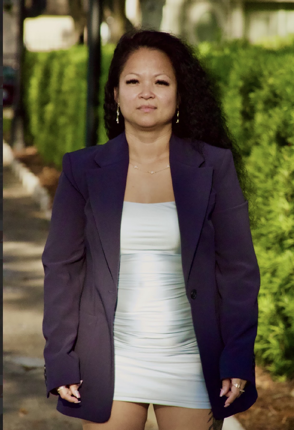 Woman with curly hair in a dark blazer and white dress outdoors, greenery in background.