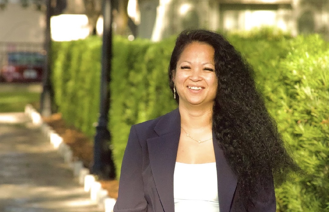 Woman with long curly hair smiling outdoors, wearing a dark blazer and white top, with green bushes in background.