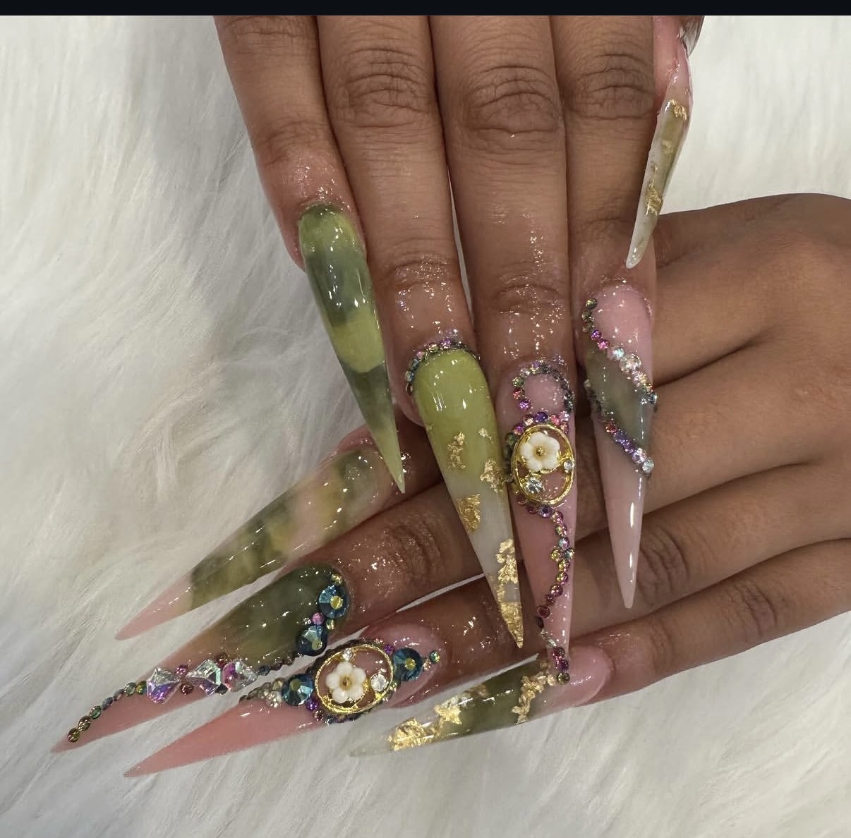 Close-up of long, decorated nails with green, pink, and gold designs, rhinestones, and floral accents, resting on a white surface.