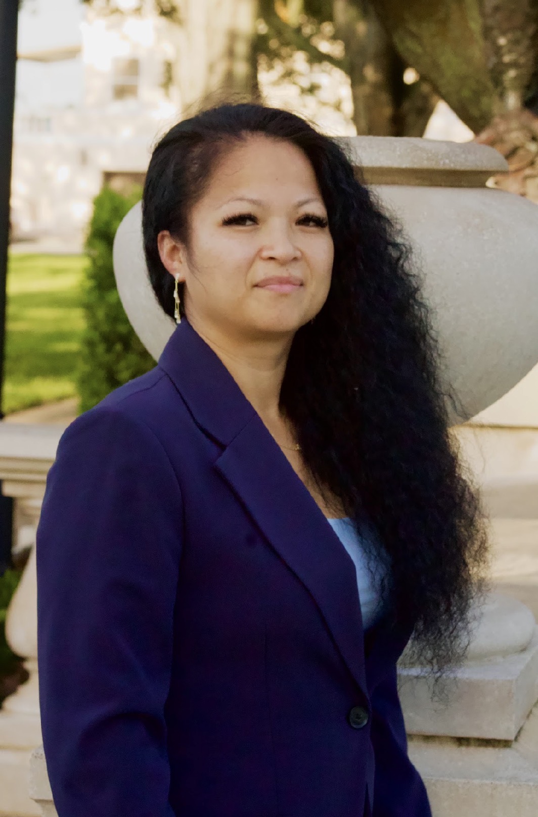 Woman with long curly hair wearing a dark blazer outdoors, with trees and a large planter in the background.