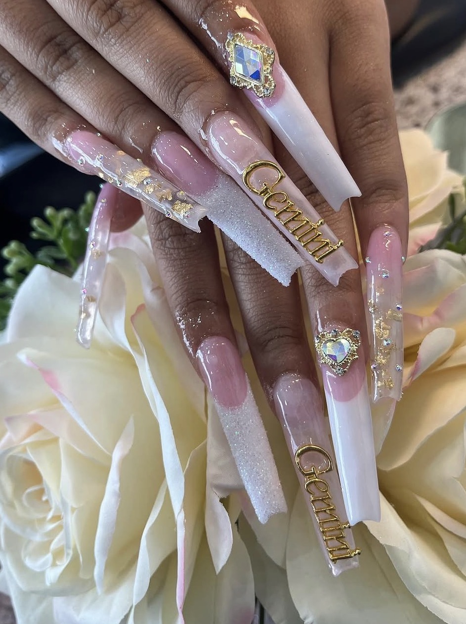Close-up of decorated long acrylic nails with gold accents and gemstones, resting on white flowers.