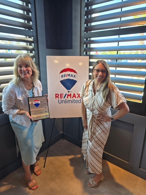 Two women stand beside a RE/MAX Unlimited sign inside a room with large window blinds, smiling at the camera.