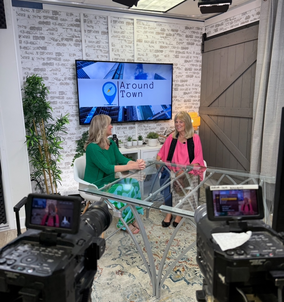 Two women sit at a glass table in a studio, talking, with a large screen behind them displaying 'Around Town'.