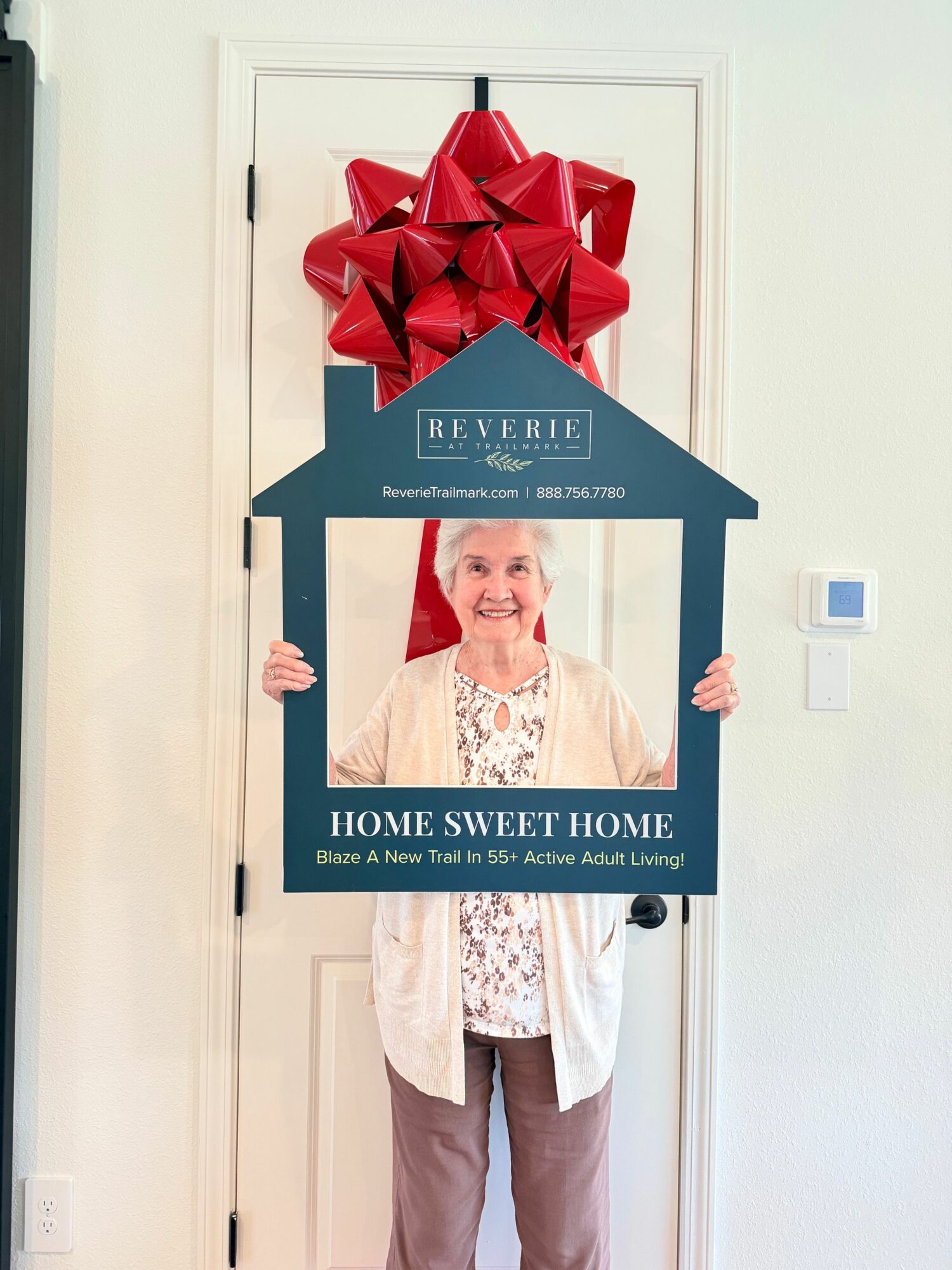Person holding a large sign with a photo and text, standing in front of a door with a red bow decoration, in a room with white walls.