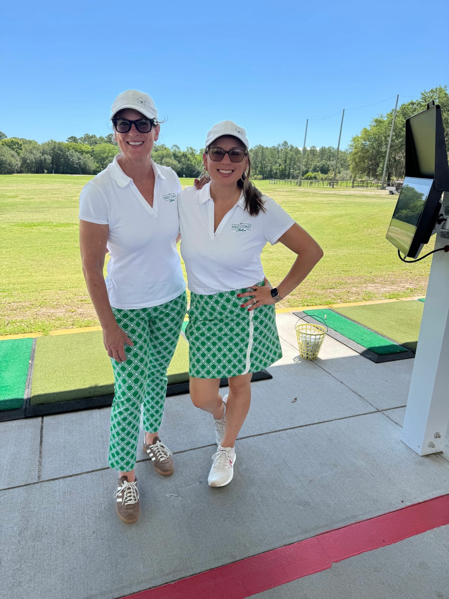 Two women standing outdoors on a sunny day, one wearing sunglasses and a white top, the other in a white shirt and green patterned pants, smiling.
