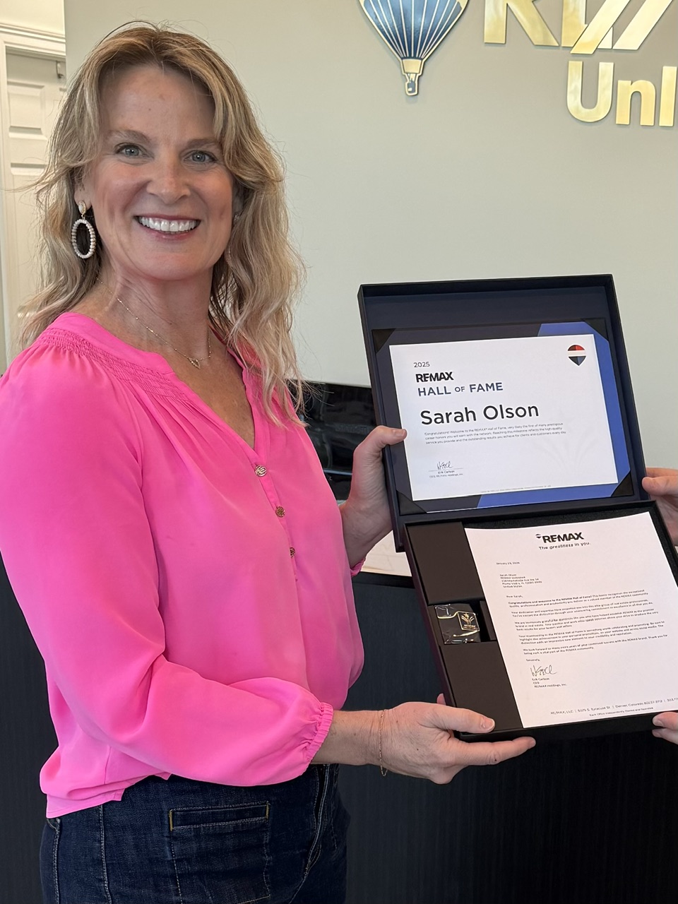 Woman with blonde hair in a pink blouse holding an award plaque, smiling, in front of a wall with a sign.
