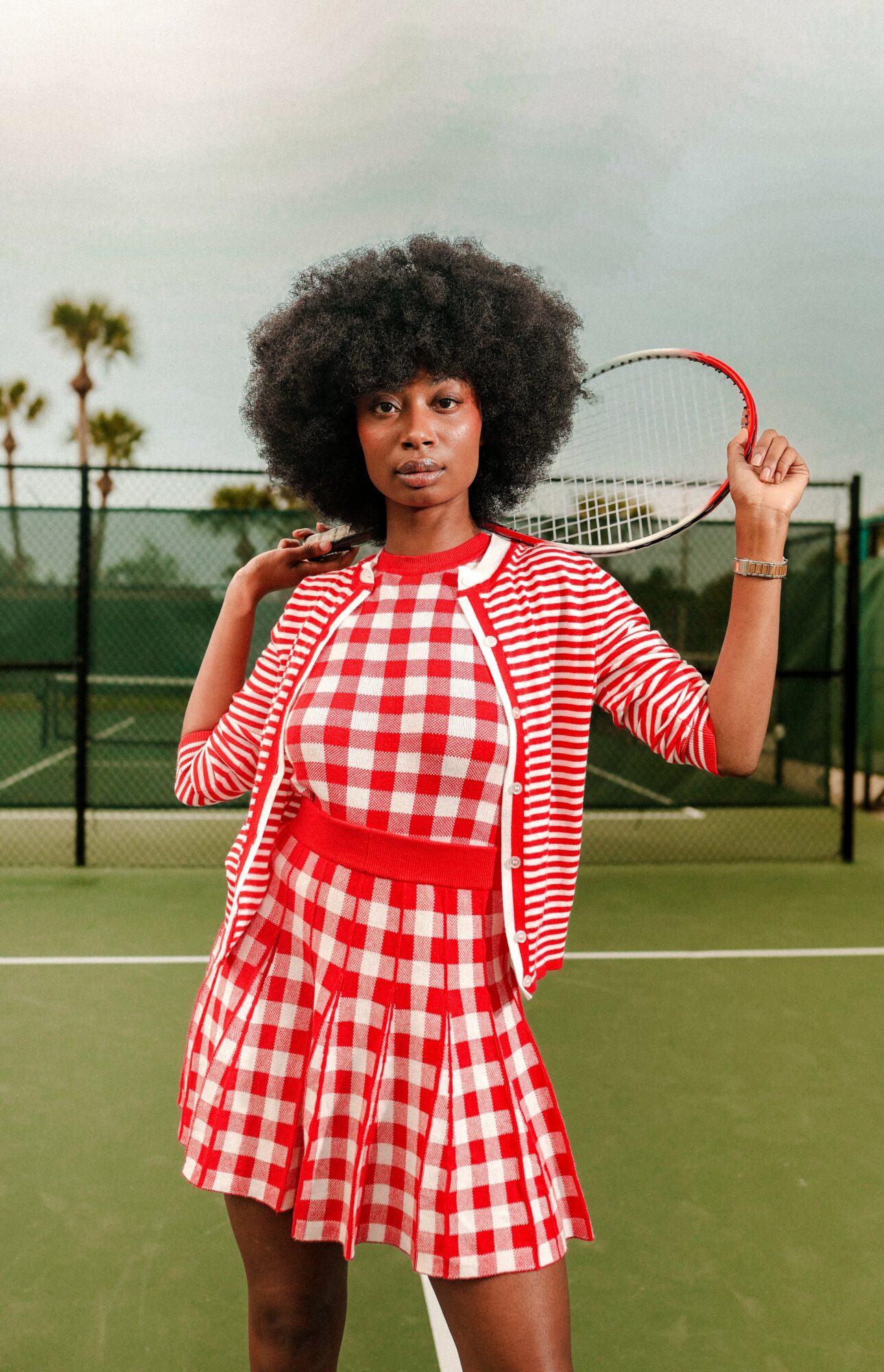 Woman with curly hair holding a tennis racket on a tennis court, wearing a red and white checkered outfit.