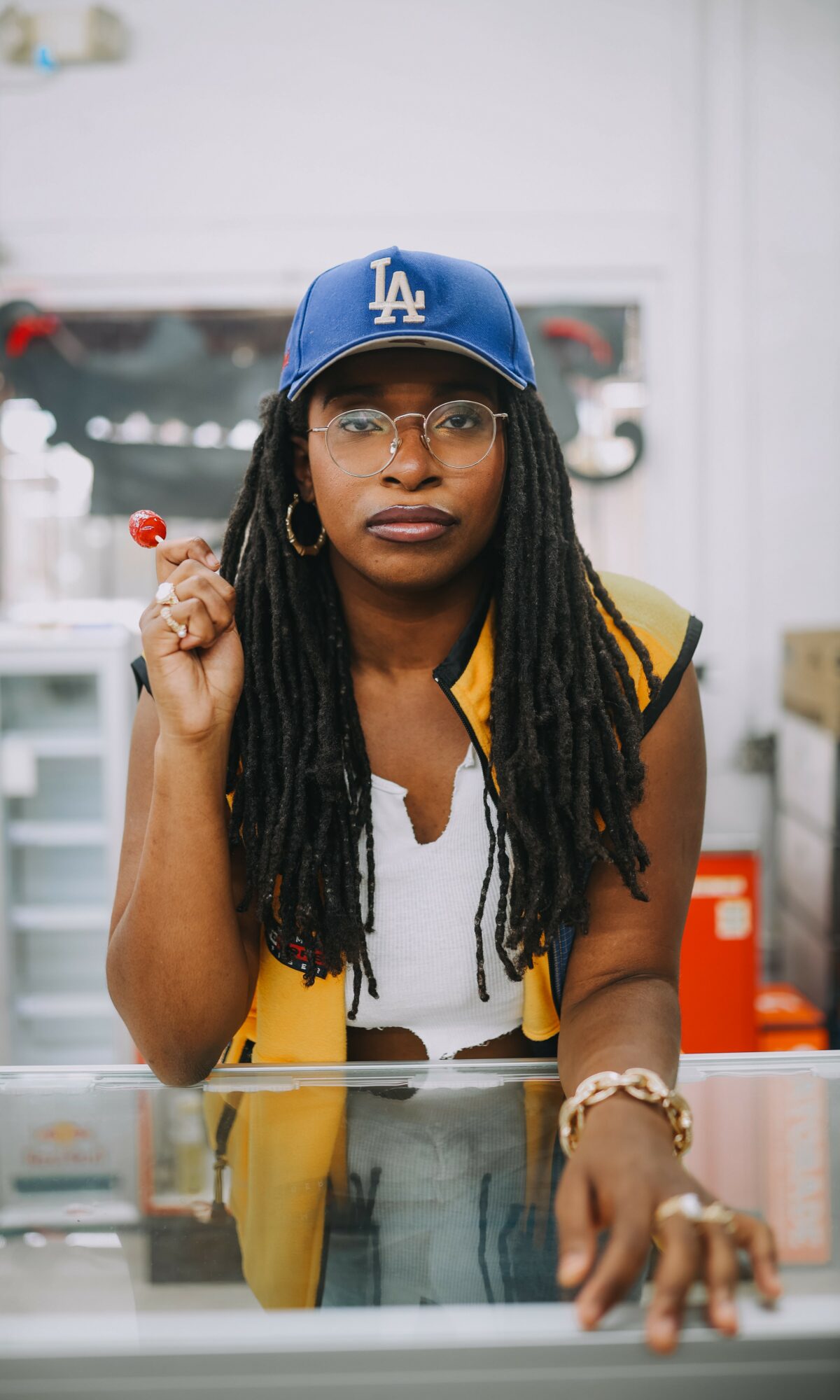 Woman with long dreadlocks wearing glasses, a blue Los Angeles Dodgers cap, and a yellow vest, holding a lollipop.