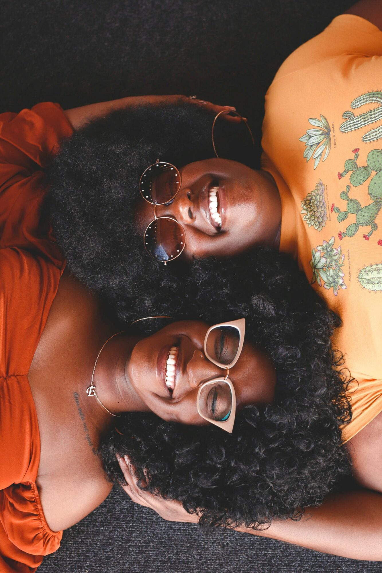 Two women with curly hair lying on dark surface, smiling, wearing sunglasses and colorful tops, with tattoos visible on one woman's arm.