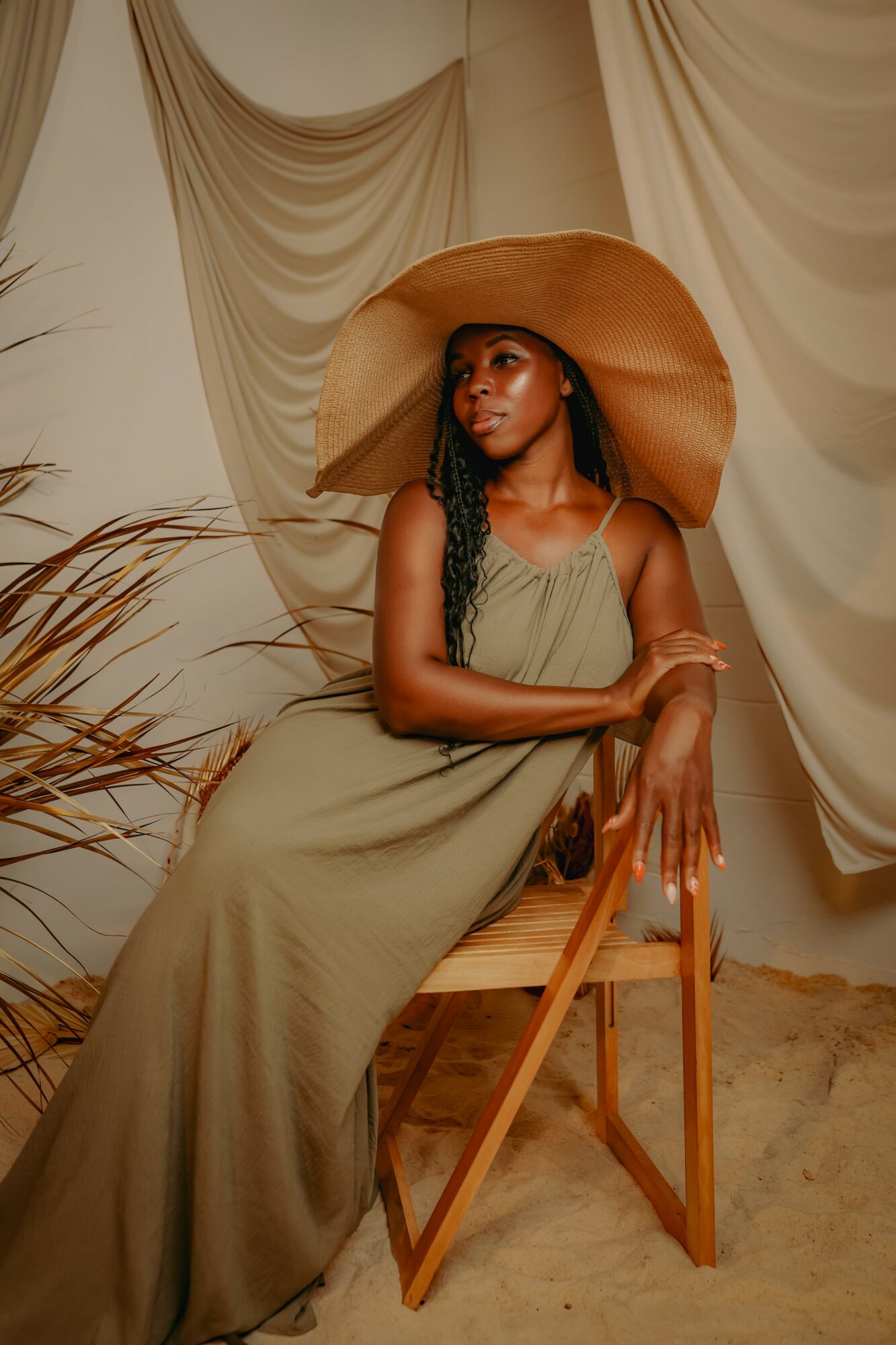 Woman in a long dress and wide-brimmed hat sitting on a wooden chair, with draped fabric background and sandy floor.
