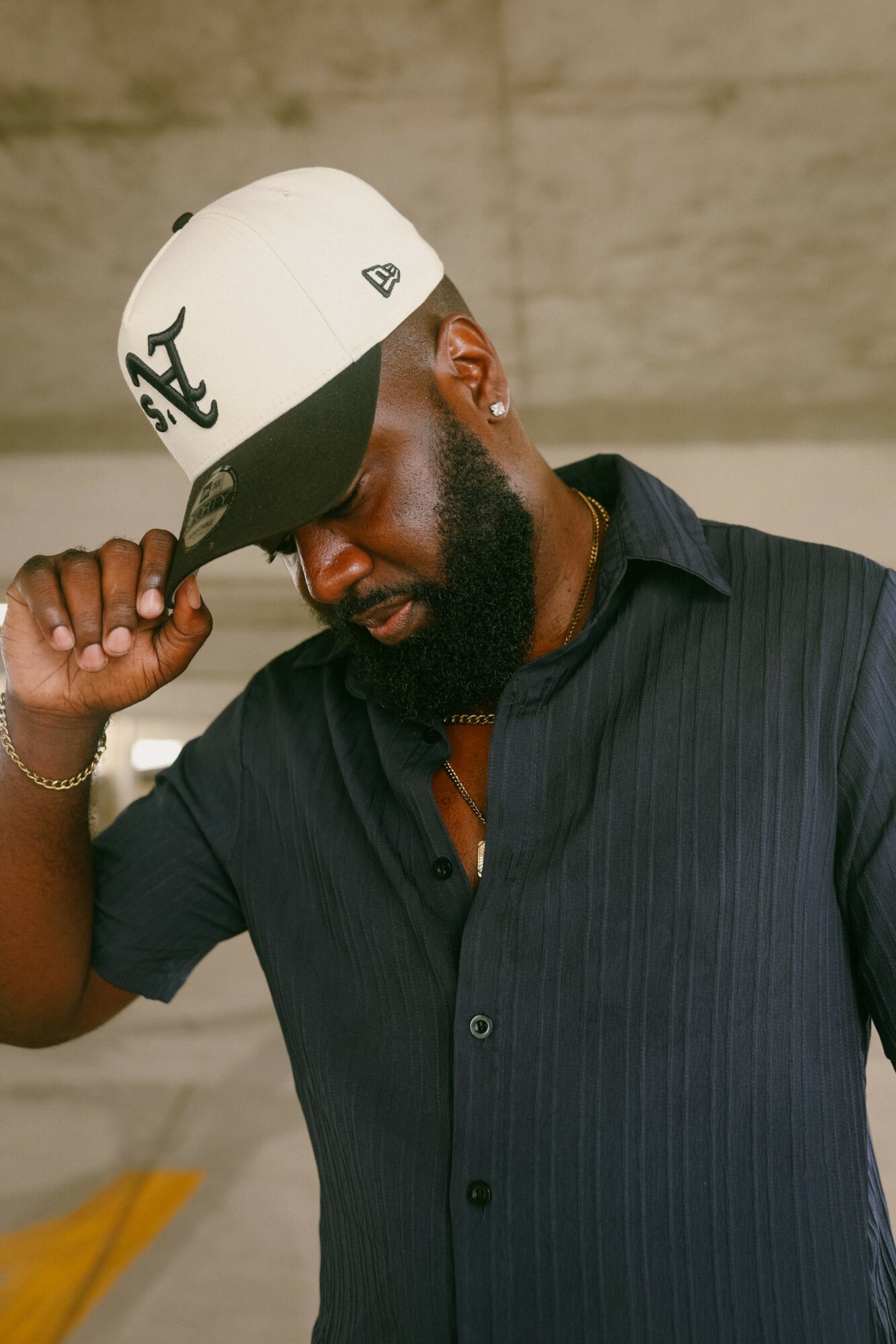 Man wearing a white cap, black shirt, and earrings, adjusting his cap with his right hand, against a neutral background.