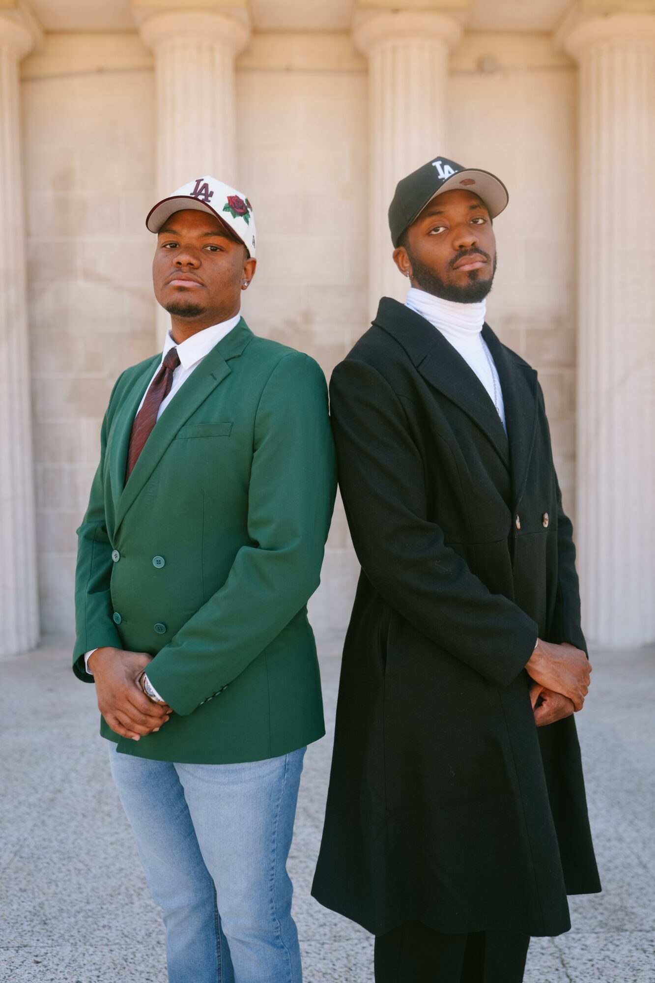 Two men stand side by side in front of large columns, dressed in formal and casual attire, looking at the camera.