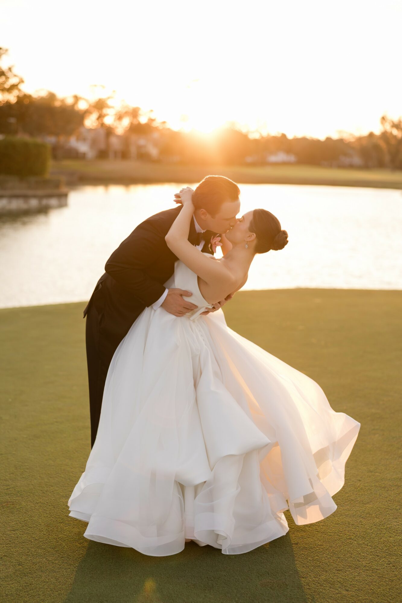 A couple in wedding attire sharing a kiss outdoors at sunset near a body of water.