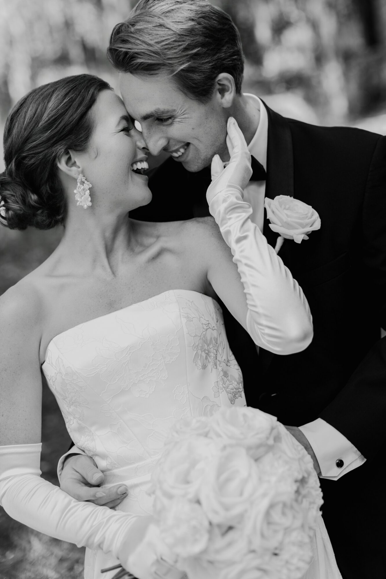 A bride and groom smile closely, touching foreheads, in wedding attire outdoors, black and white photo.