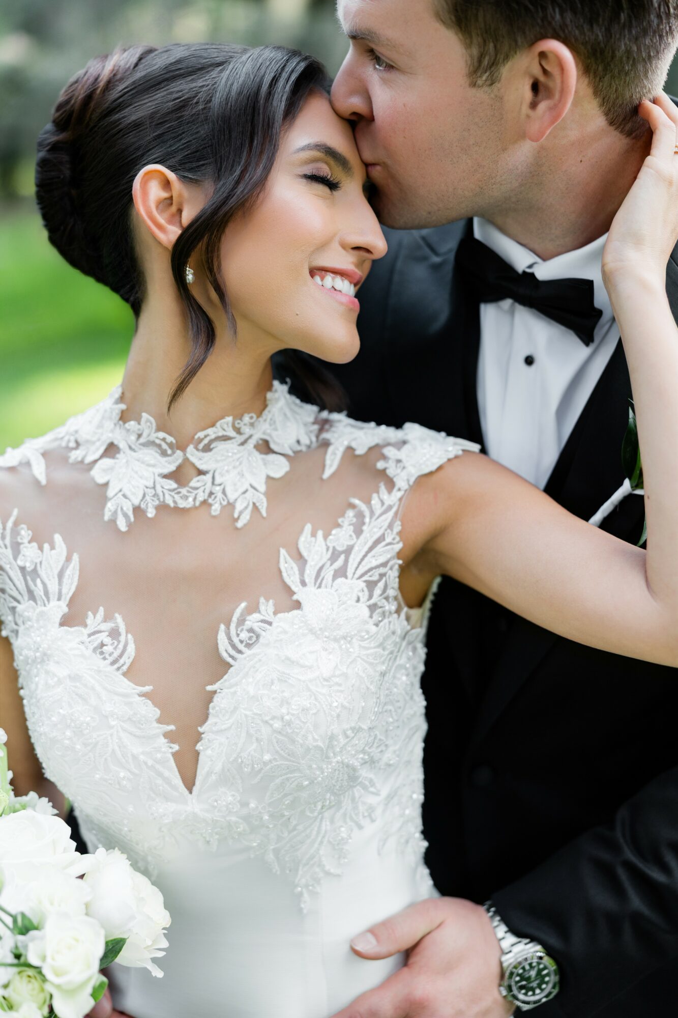 A bride and groom smile and embrace outdoors, with the groom kissing the bride's forehead, both dressed in wedding attire.