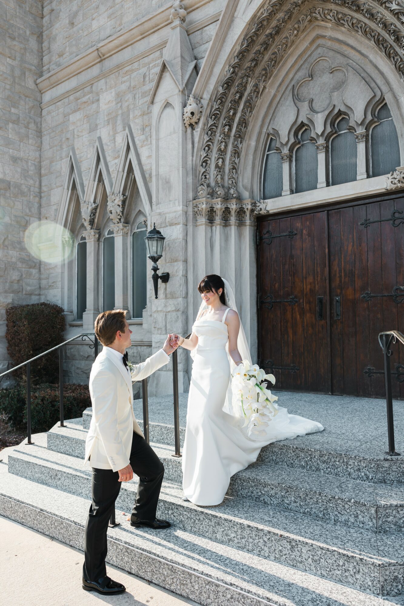 A bride in a white dress and a groom in a white suit exchange a handshake on church steps outside a stone building.