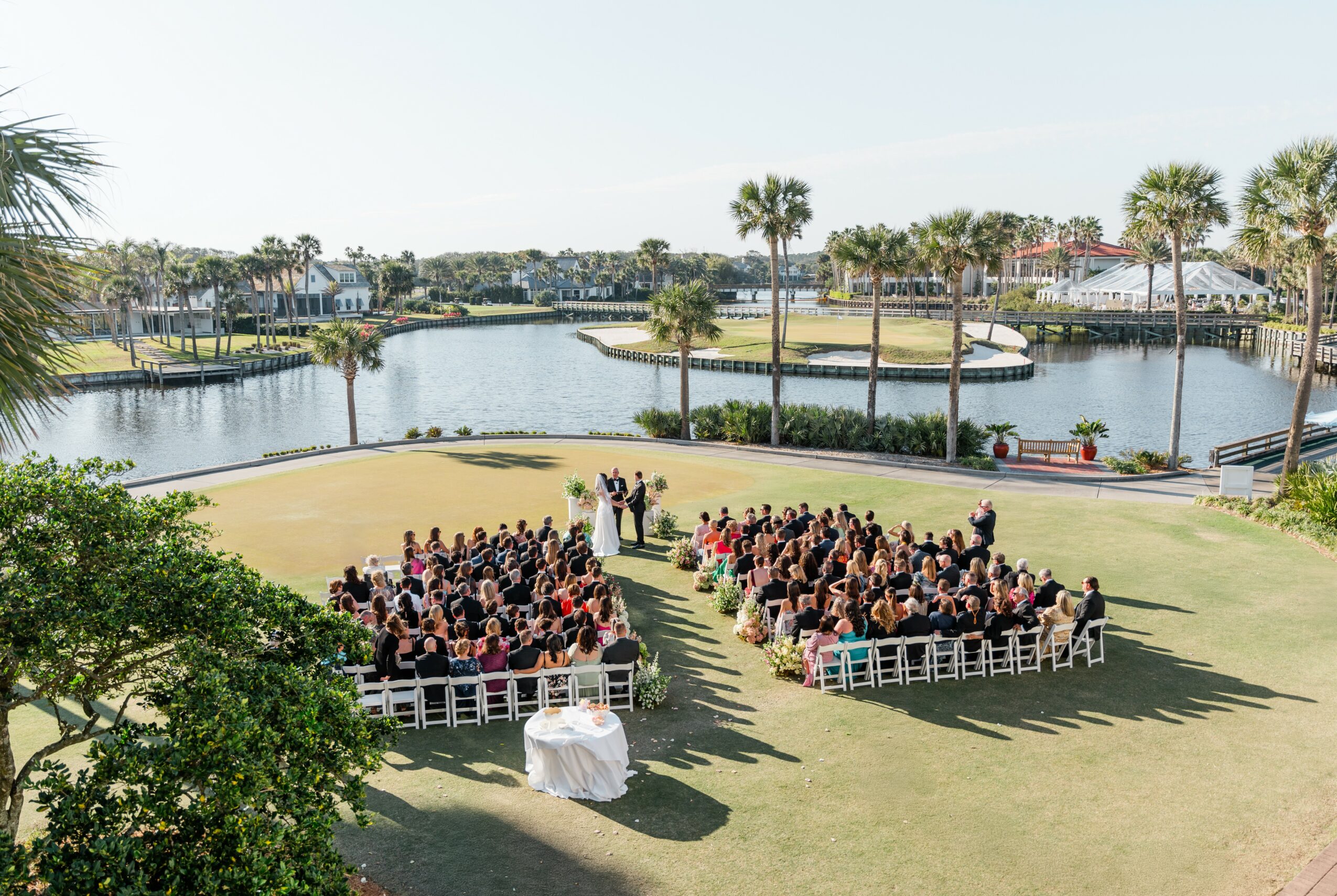 Outdoor wedding ceremony with guests seated on chairs facing a couple, water and palm trees in background.