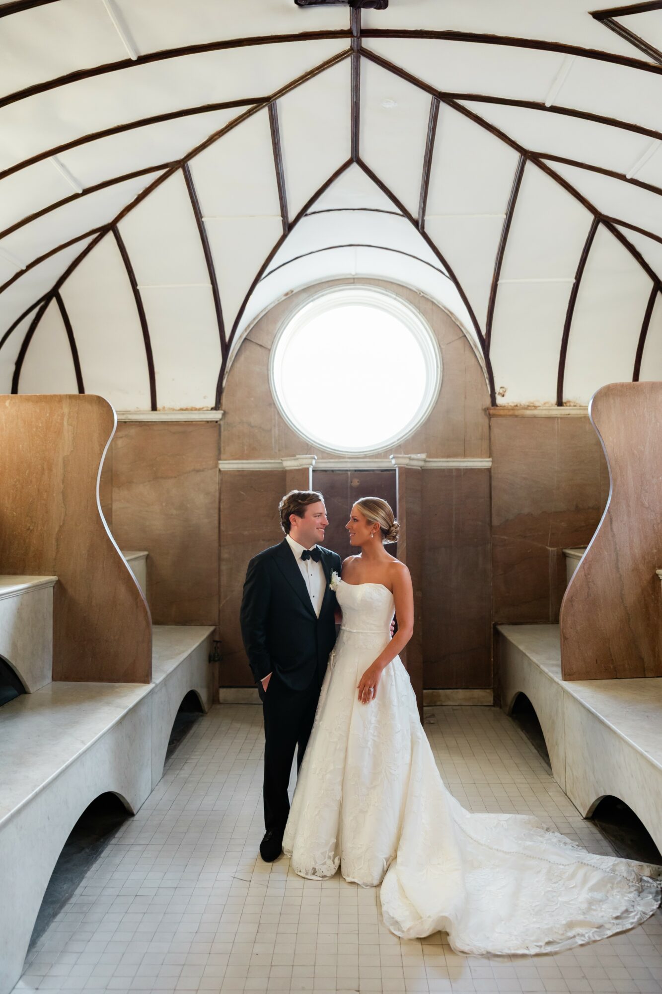 Bride and groom stand inside a church with a circular window and curved wooden architecture, facing each other.
