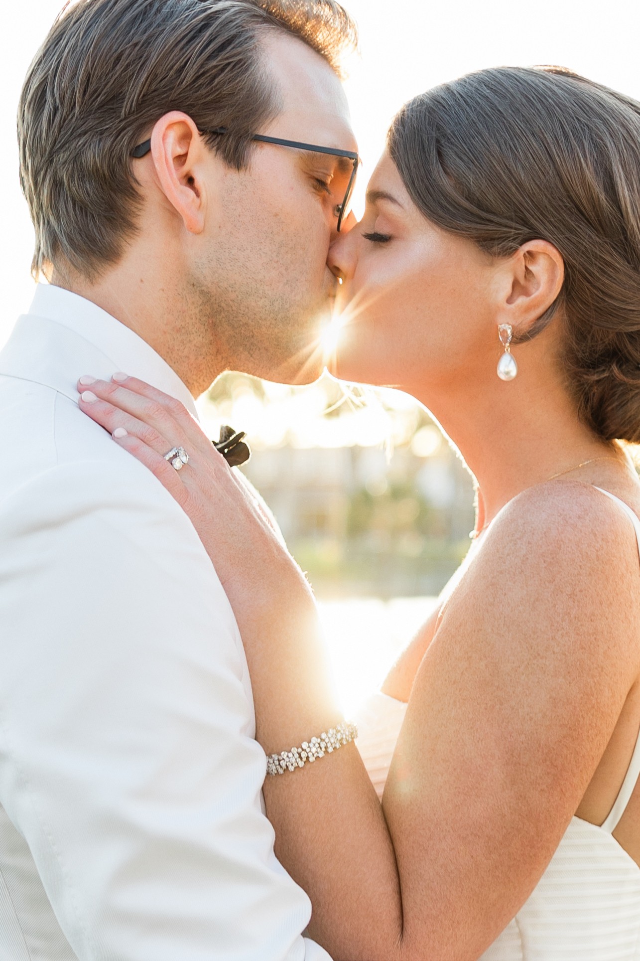 A couple kissing outdoors with sunlight shining between them, dressed elegantly, with the woman wearing earrings and a bracelet.