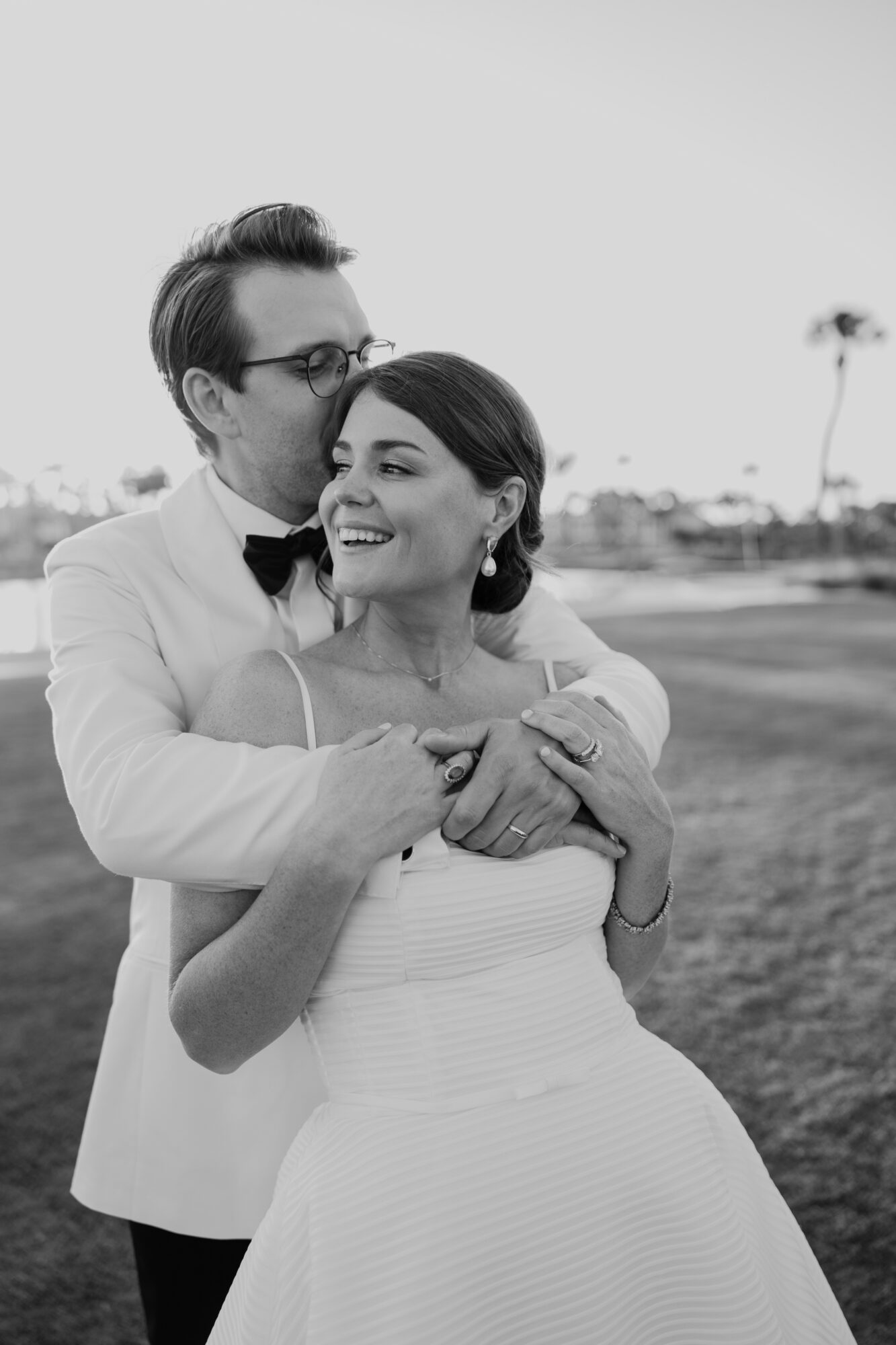 A couple in formal attire embracing outdoors, smiling, with a blurred background of trees and sky.