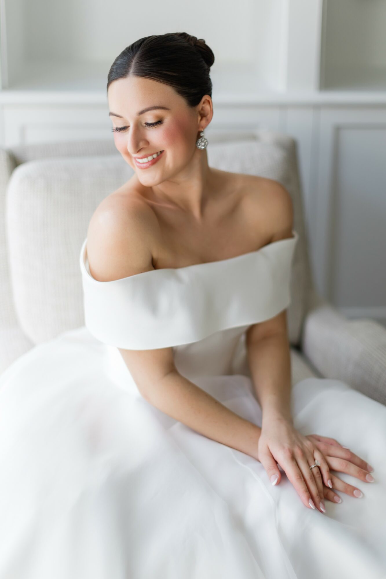 Smiling woman in white dress sitting on a sofa with hands on lap, indoors, soft lighting.