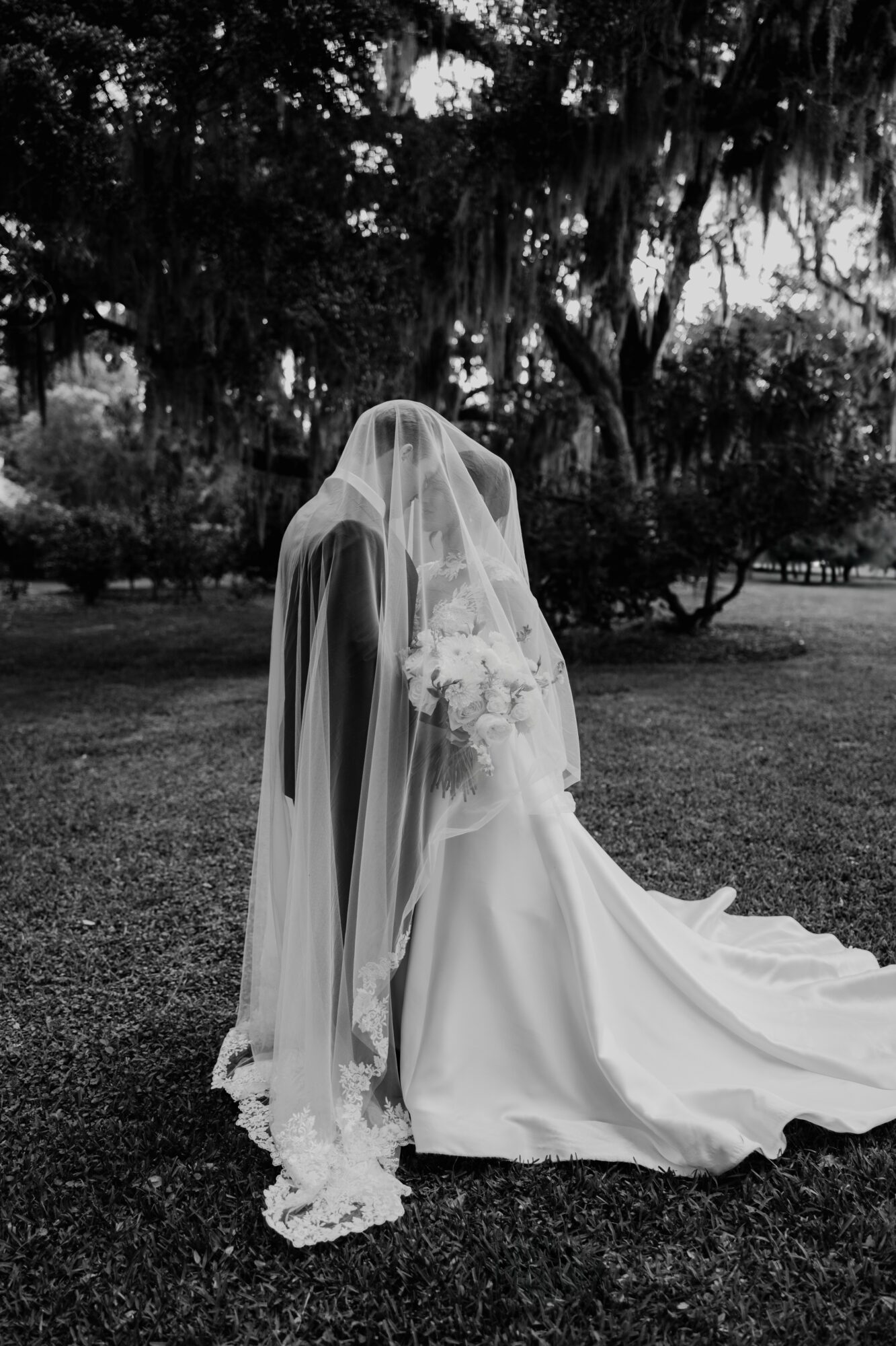 Bride in wedding dress with veil kneeling outdoors, holding bouquet, trees in background.