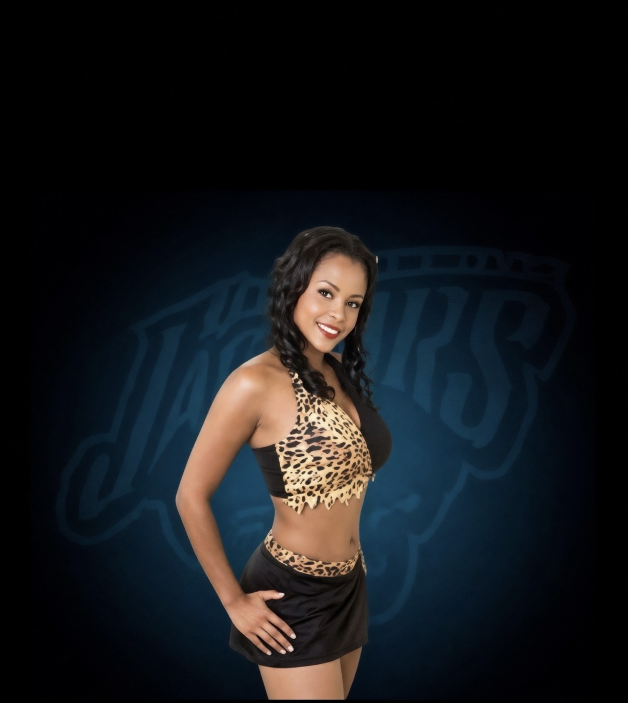 Young woman with long dark hair smiling, wearing a leopard print top and black shorts, standing against a dark background with a logo.