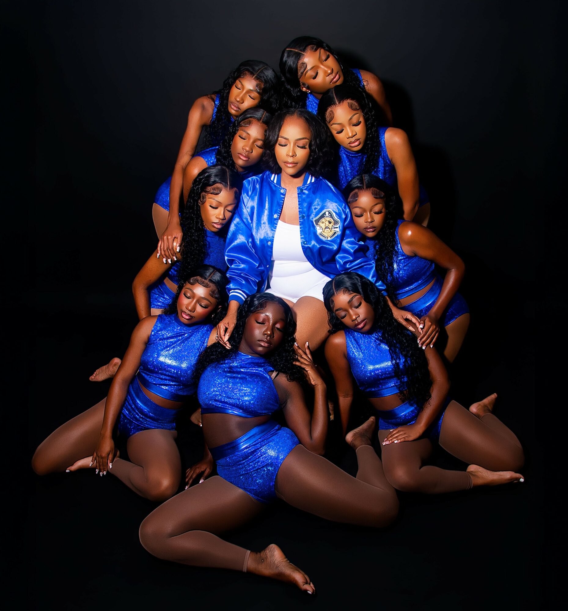 Group of nine young women in blue outfits, posing together against a black background.