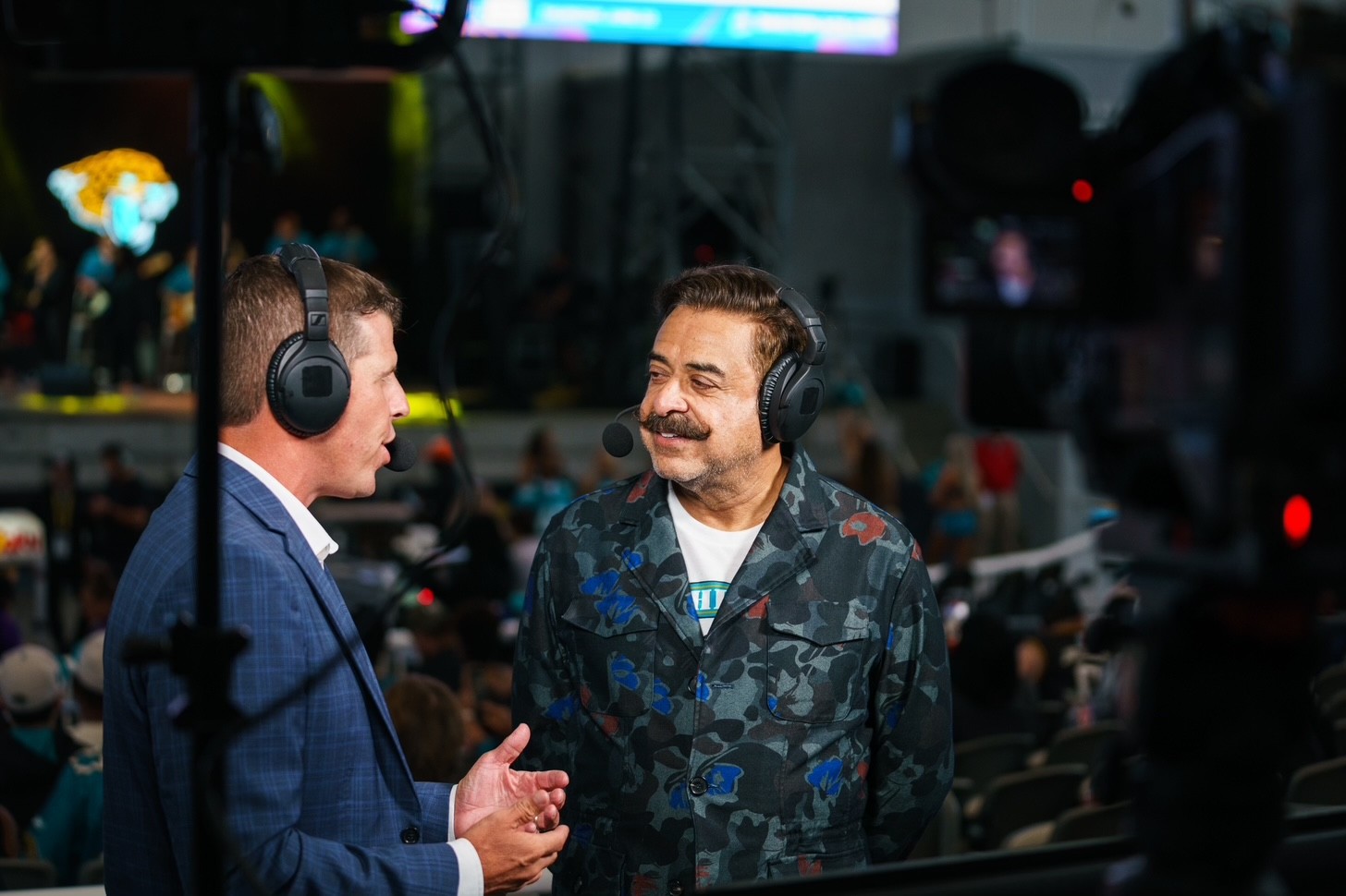 Two men wearing headphones and talking in a studio or control room, with equipment and screens in the background.