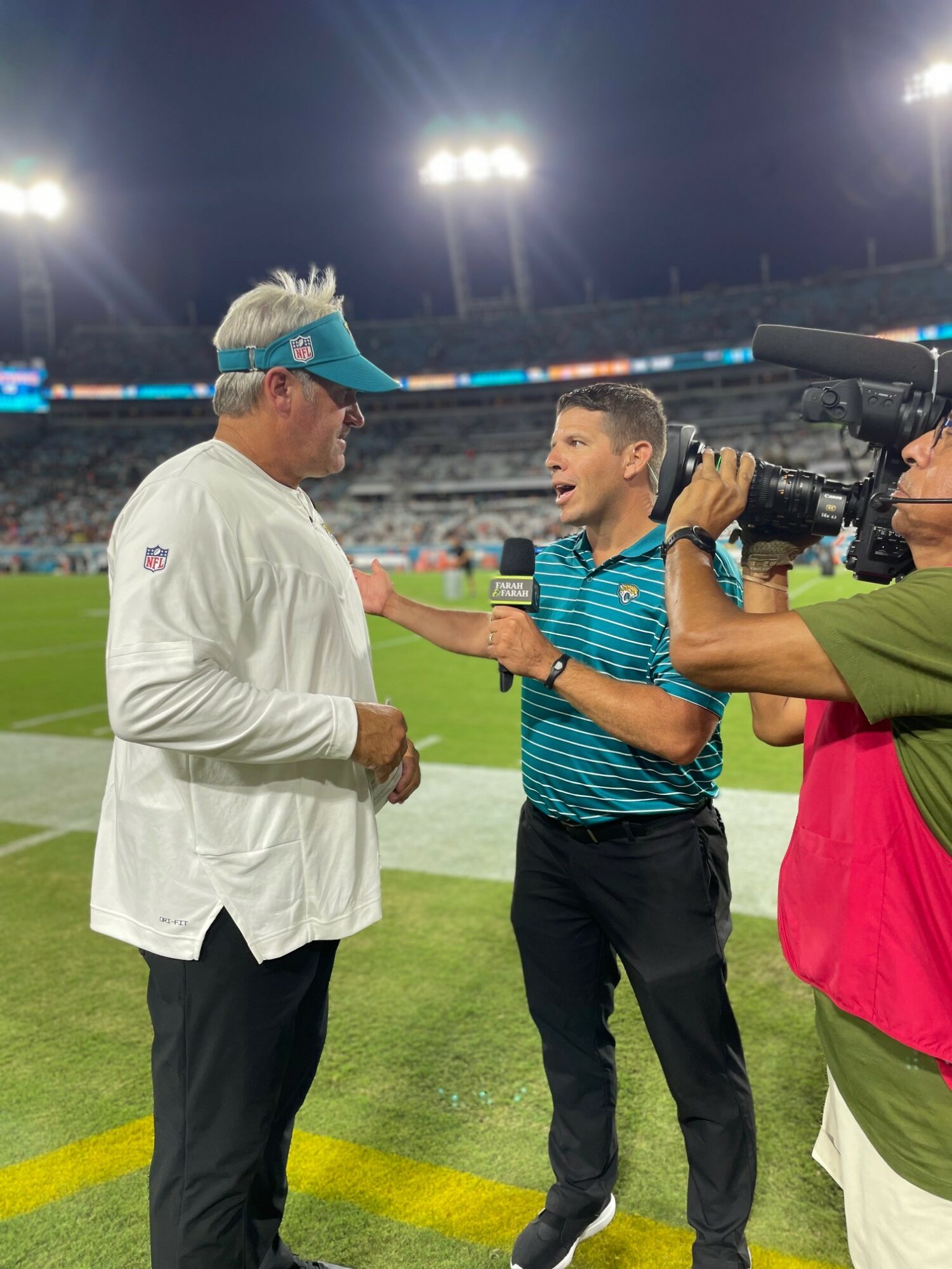 Two men and a cameraman on a sports field at night, with stadium lights overhead, one man wearing a white shirt and cap, the other in a striped shirt, talking animatedly.