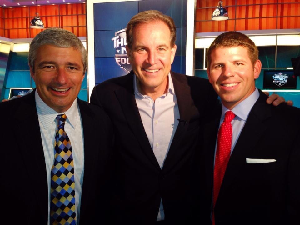 Three men in suits smiling, standing in a studio with bright lights and screens behind them.
