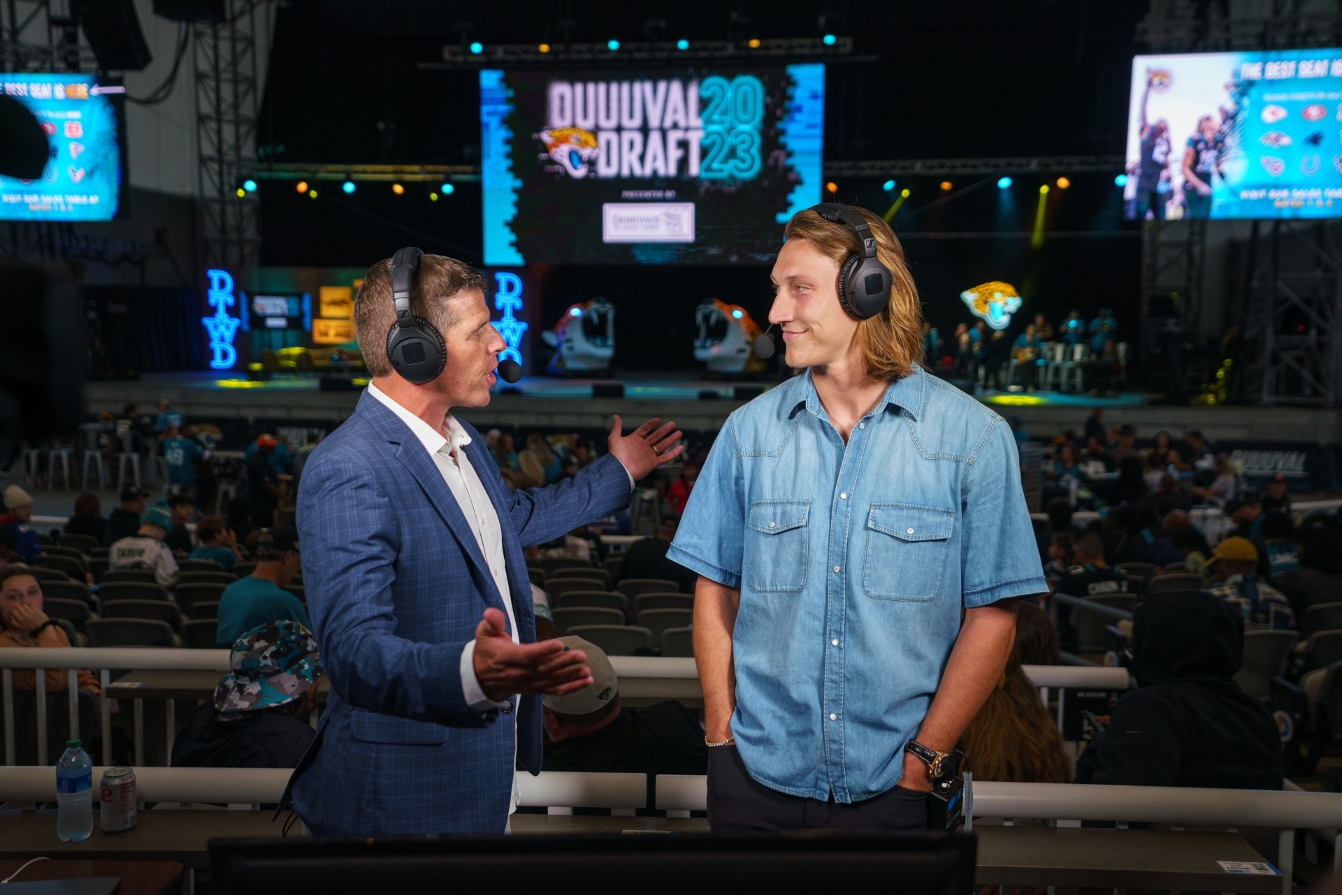 Two men with headsets talking at an esports event, large screens and audience in background.