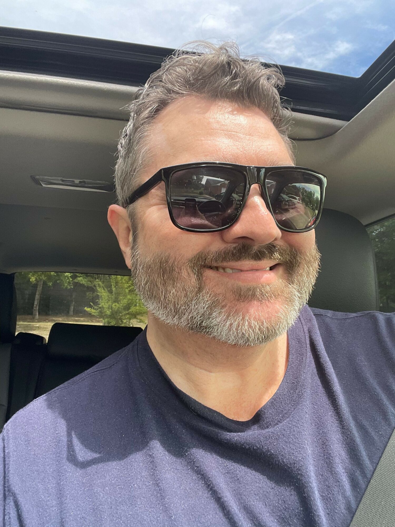 Man with sunglasses and gray beard sitting in a vehicle, wearing a dark shirt, with a view of the sky outside.