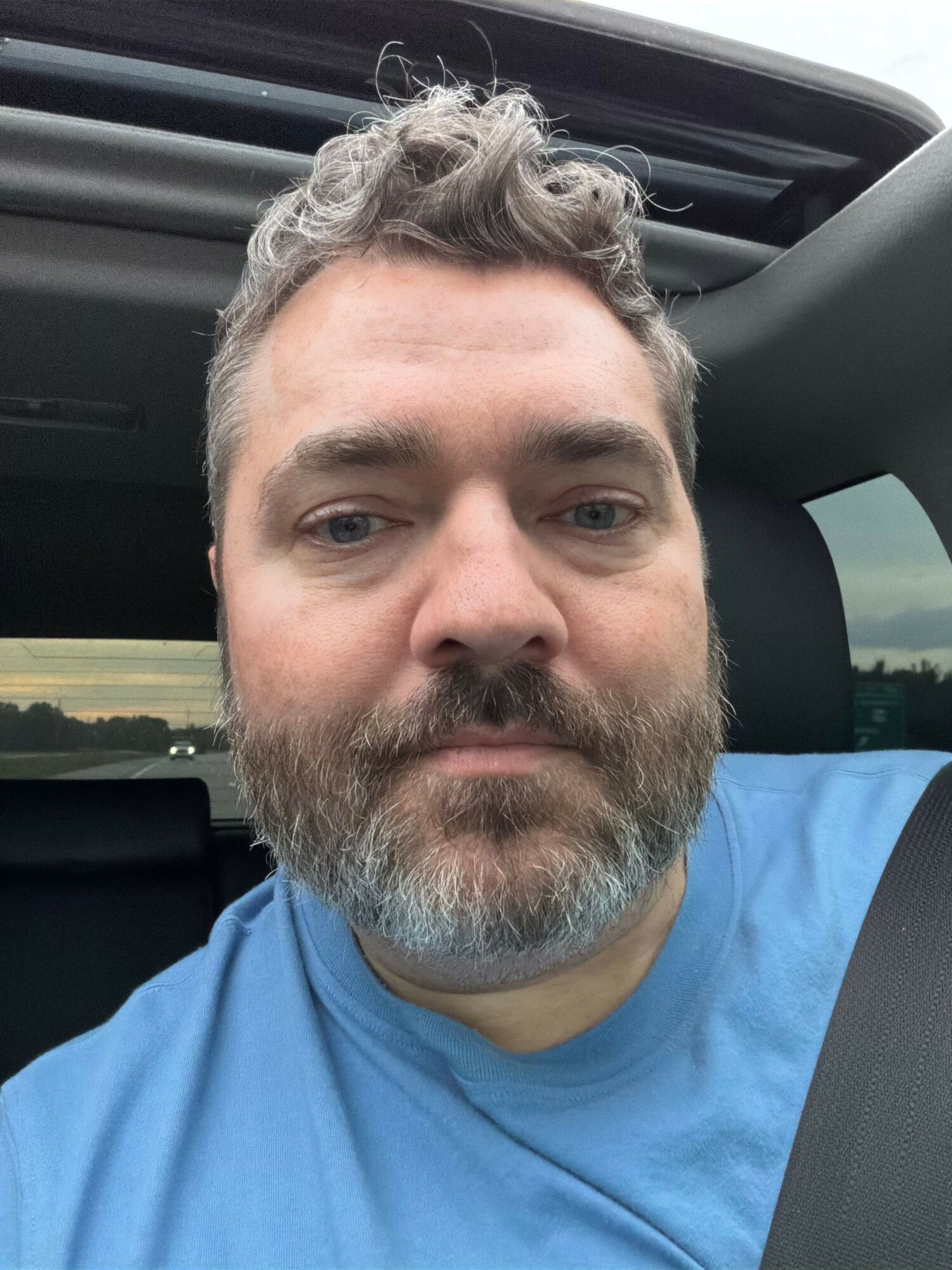 Man with gray hair and beard sitting in a vehicle, wearing a blue shirt, looking at the camera.