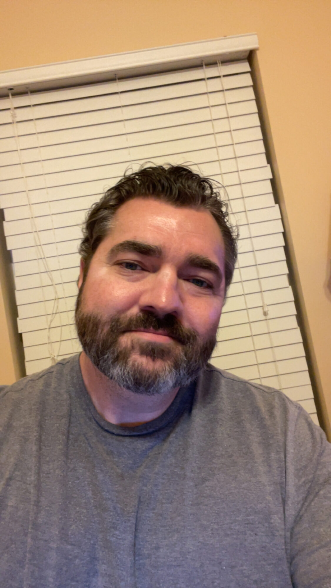 Man with dark hair and beard wearing a gray shirt, smiling, in front of closed blinds and a beige wall.