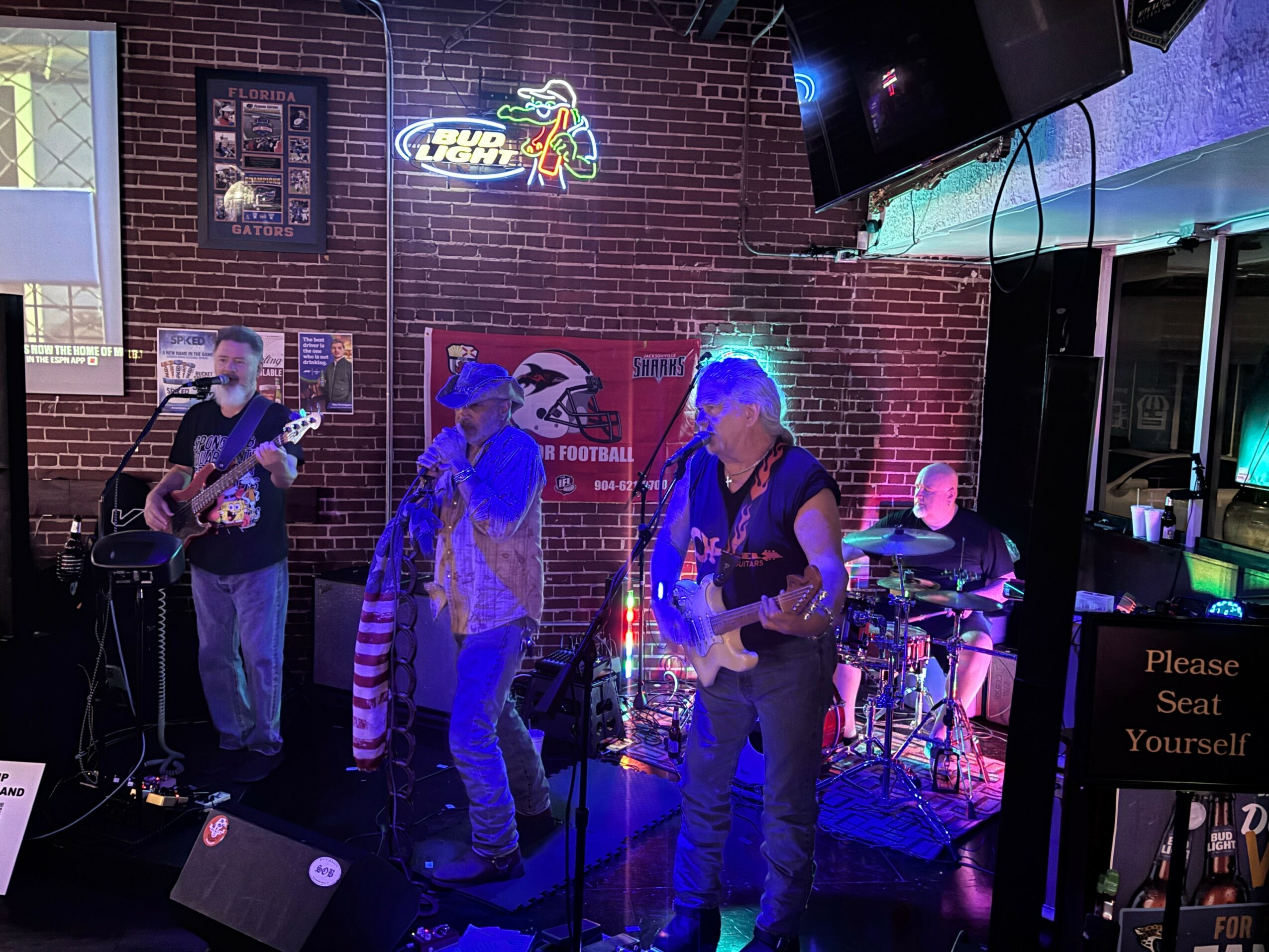 Four musicians perform on stage with brick wall background, neon signs, and musical equipment, in a dimly lit venue.