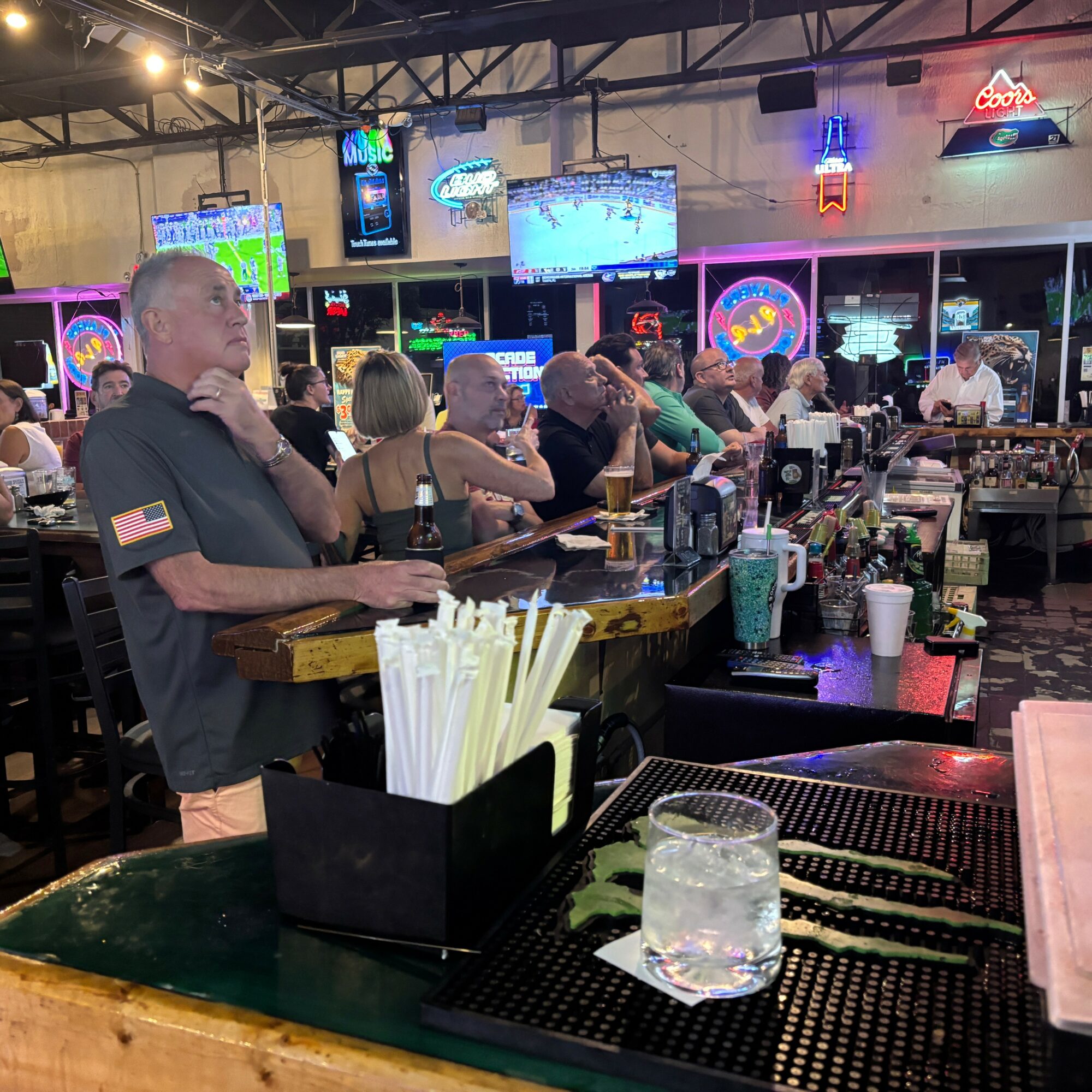 People sitting at a bar with drinks, neon signs, and multiple TV screens in the background.