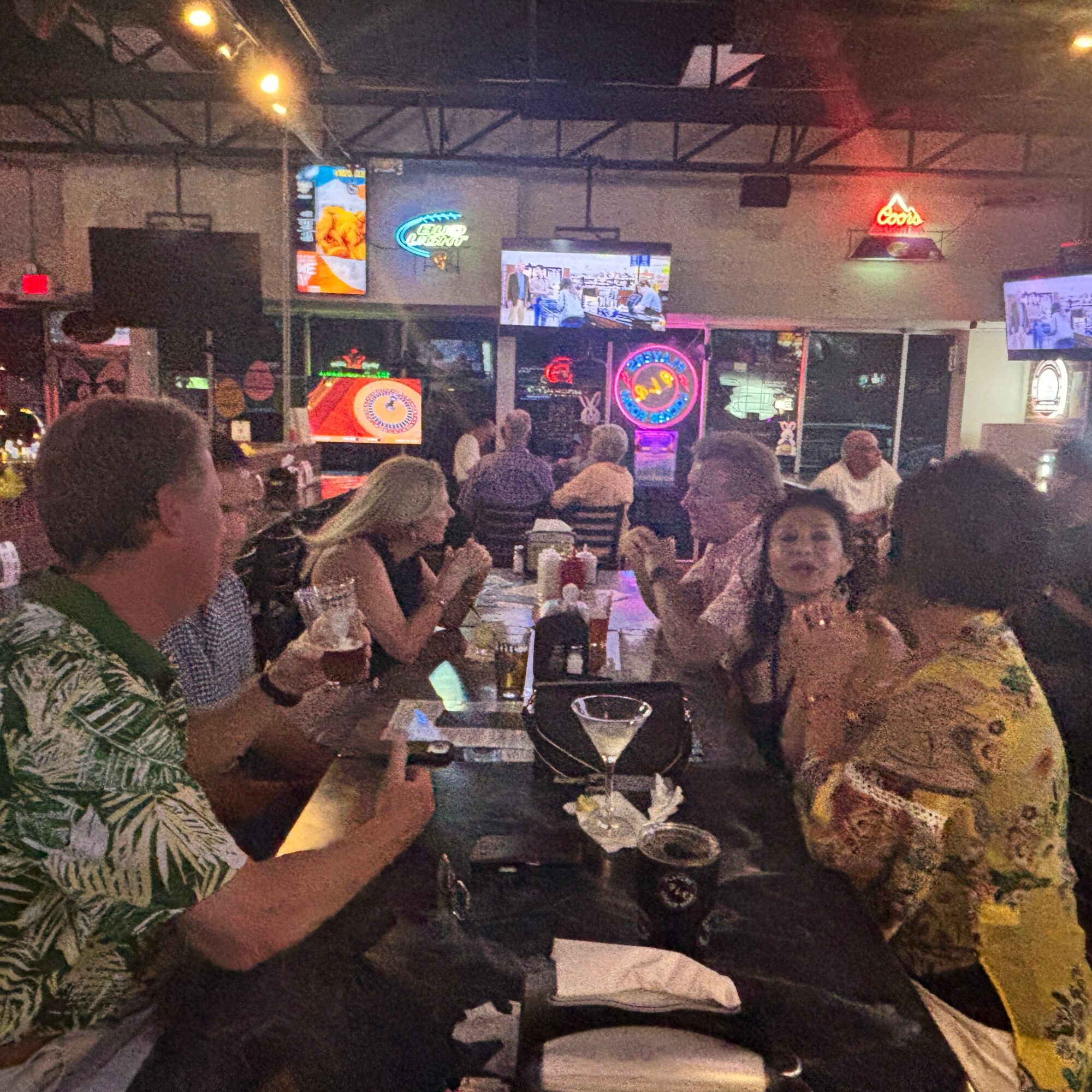 People sitting at a bar or restaurant, socializing, with multiple TV screens and neon signs in the background.