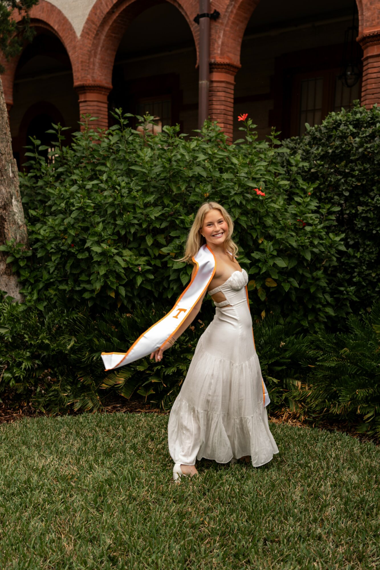 Young woman in a white dress standing outdoors in front of green bushes, smiling, with a building in the background.